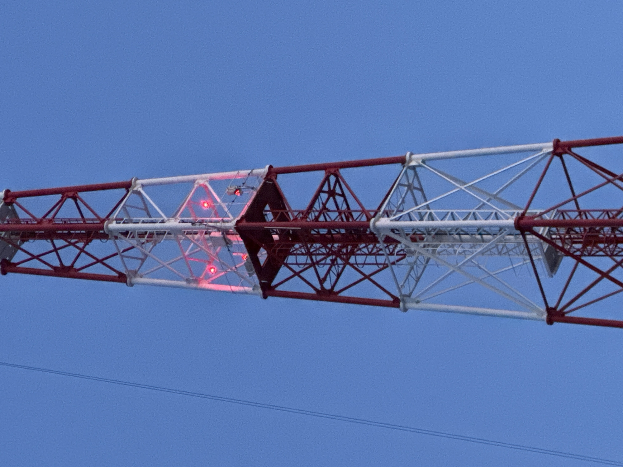 The image depicts a section of a lattice tower structure against a clear blue sky. The tower is painted in alternating sections of red and white. There are red lights visible within the white section. The location is unspecified and there's no provided context to establish this is in Chaikovsky, Russia. No people, actions or interactions are visible.