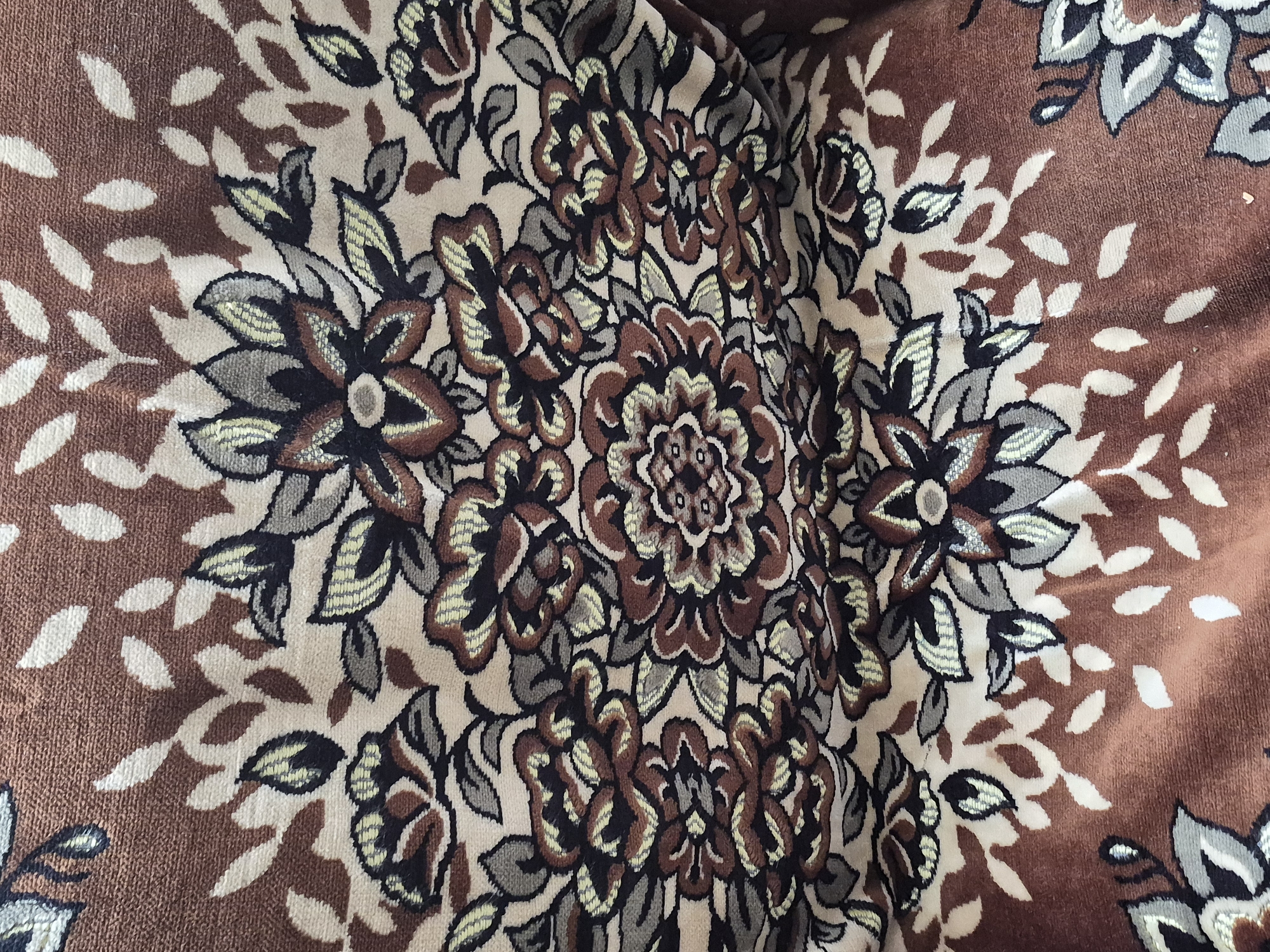 The image shows a close-up of a brown fabric with a floral pattern. The pattern consists of stylized flowers and leaves in shades of brown, beige, black, and grey. The fabric appears to be textured, possibly a rug or upholstery. There are no people or discernible environmental features visible that would indicate a specific location in Vanadzor, Armenia. The image focuses solely on the textile design.