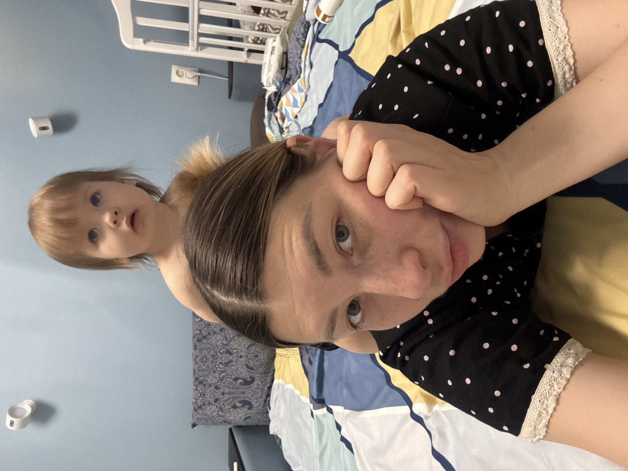 A woman in a black and white polka dot garment lies on a bed covered with a blue, yellow, and white patterned bedspread. A young child with blonde hair and bangs stands behind her. The room is painted blue. A white crib is visible in the background. The location is not definitively discernible as Moscow, Russia, based on the image alone.