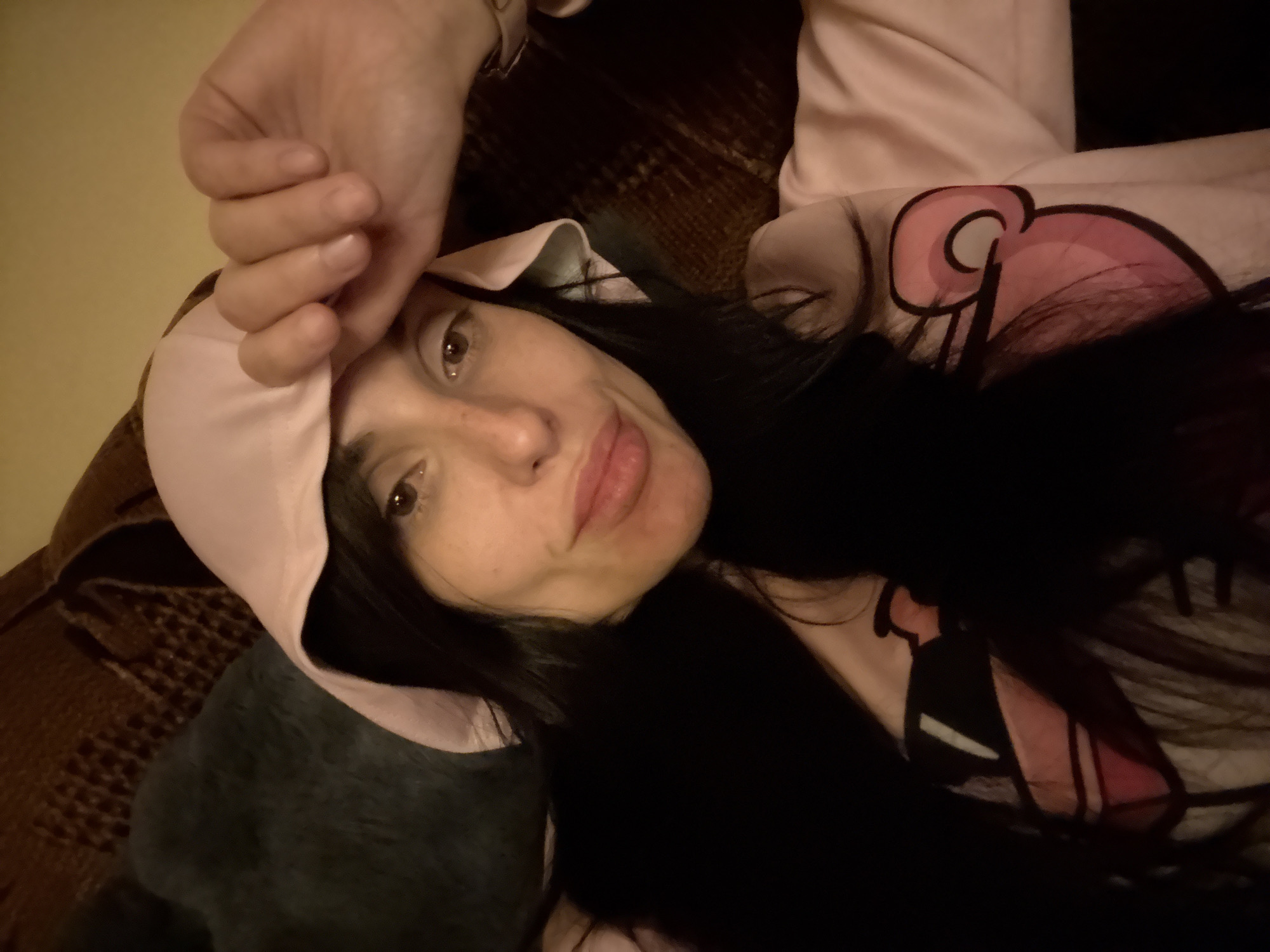 An indoor scene in Pleven, Bulgaria, features a female individual posing for the camera. The subject has long, dark hair extending over her shoulders and chest. She is wearing a light pink hooded garment, with the hood pulled up, and a matching long-sleeved top that displays a pink, black, and white cartoon graphic on the right chest area. Her right hand is raised, with fingers resting on her forehead, partially obscuring the upper portion of her face and the hood. She has dark eyes with visible white eyeliner on the lower waterline and full lips. Her gaze is directed towards the camera. The background is dimly lit and out of focus, revealing a dark, plush object to the left and a dark brown, patterned upholstered surface behind the subject.