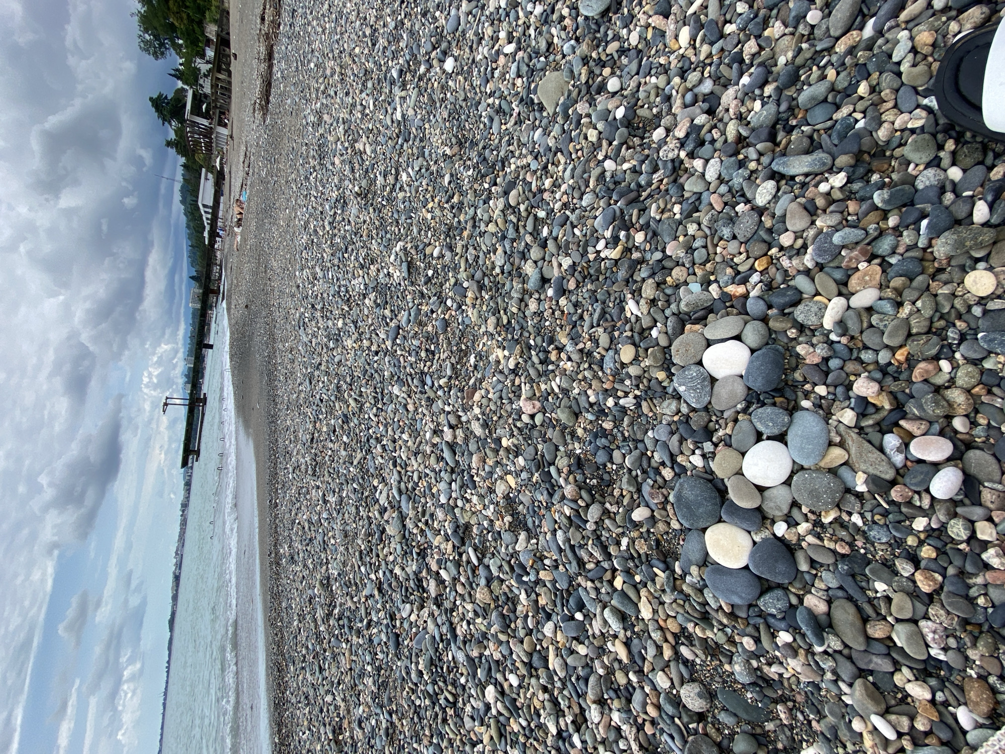 The image depicts a pebble beach in Sokhumi, Abkhazia, under a cloudy sky. In the foreground, several smooth, light-colored pebbles are arranged in the shape of a heart. The beach is densely covered with a variety of gray, black, and white pebbles. In the middle ground, the sea meets the shore with small waves. A pier extends into the water on the left side of the image, and buildings are visible along the coastline in the distance. Some people can be seen sunbathing on the beach further down. The overall scene is one of a coastal landscape on a potentially overcast day.