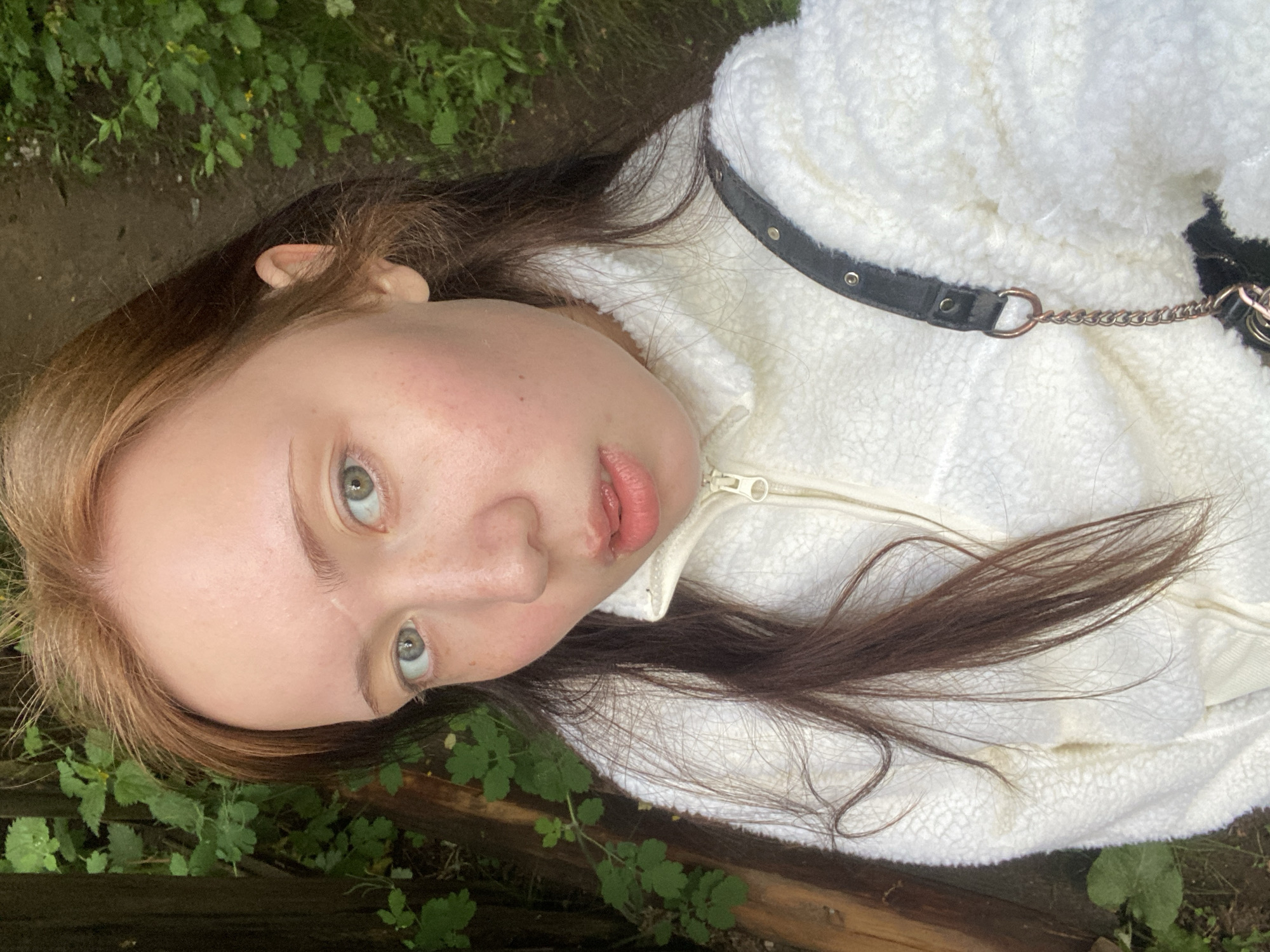 A young person lies on a patch of ground overgrown with green foliage, possibly near a wooden structure. They are wearing a white, textured jacket with a black strap and chain visible. The individual has brown hair with lighter streaks and fair skin. Their eyes are open, looking upwards. The location appears to be outdoors. The city is not discernible from the image.