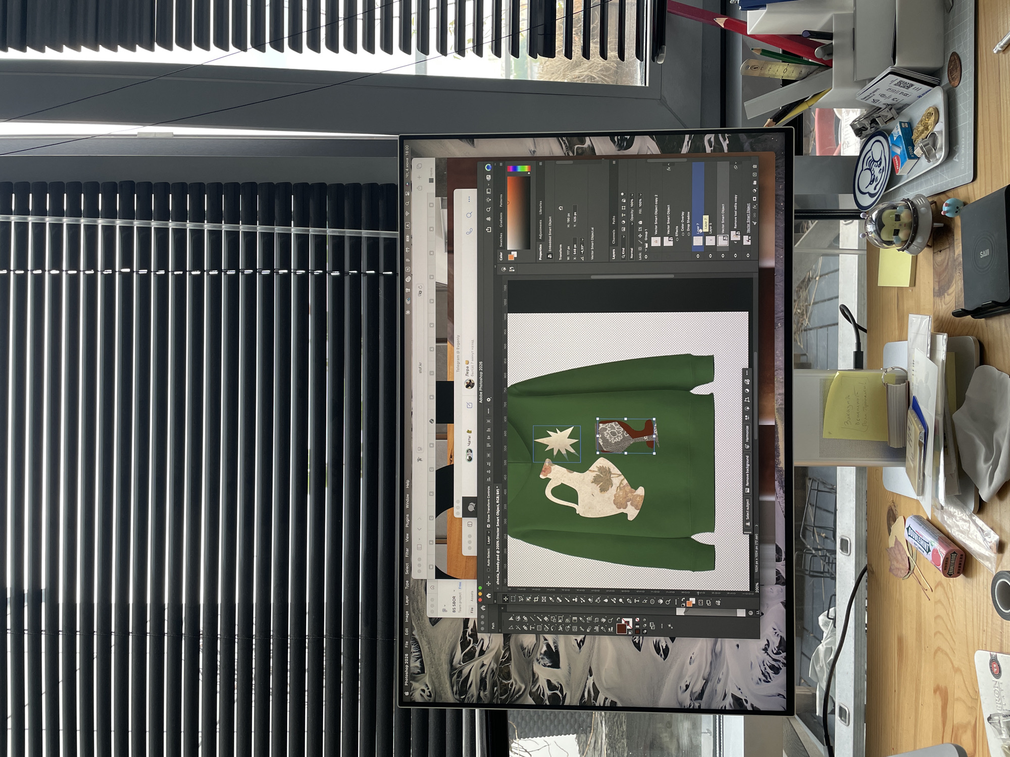 The image captures an overhead view of a creative workspace in Saint Petersburg, Russia, featuring a computer monitor displaying design software and a cluttered wooden desk. The scene is illuminated by natural light filtering through window blinds.
On the large monitor, Adobe Photoshop 2026 is open, showing a design in progress for a green hoodie. The hoodie features a white star, a patterned brown shape, and an ornate jug with floral patterns. The Photoshop interface is clearly visible, displaying various panels such as Layers, Properties, Color, and tools. Several browser tabs are open behind Photoshop, including one for "Telegram @ Evgeniy" with a message from "Лера" indicating she was "7 минут назад" (7 minutes ago). Another tab shows "stof.kr". The system clock on the monitor indicates "Чт, 4 июня 18:00" (Thursday, June 4th, 6:00 PM), suggesting it's late afternoon or early evening.
The wooden desk to the right of the monitor is covered with office supplies and personal items. A white desk organizer holds an assortment of pencils (including a prominent red one), pens, and a ruler. Next to it, a small tray contains coins, badges (one with "1000"), and a small blue figurine. A yellow sticky note with handwritten text "Заказать", "Вернемся", and "Поли Пропалки!" is visible. Other items include a pack of Doublemint gum, dried leaves, a roll of black tape, a black wired mouse, a black smartphone on a SAIJI brand stand, and a coaster with "BRONX..." branding.
The background shows white vertical window blinds, partially open, revealing daylight and suggesting an outdoor view beyond. The overall impression is that of an active digital design environment.
