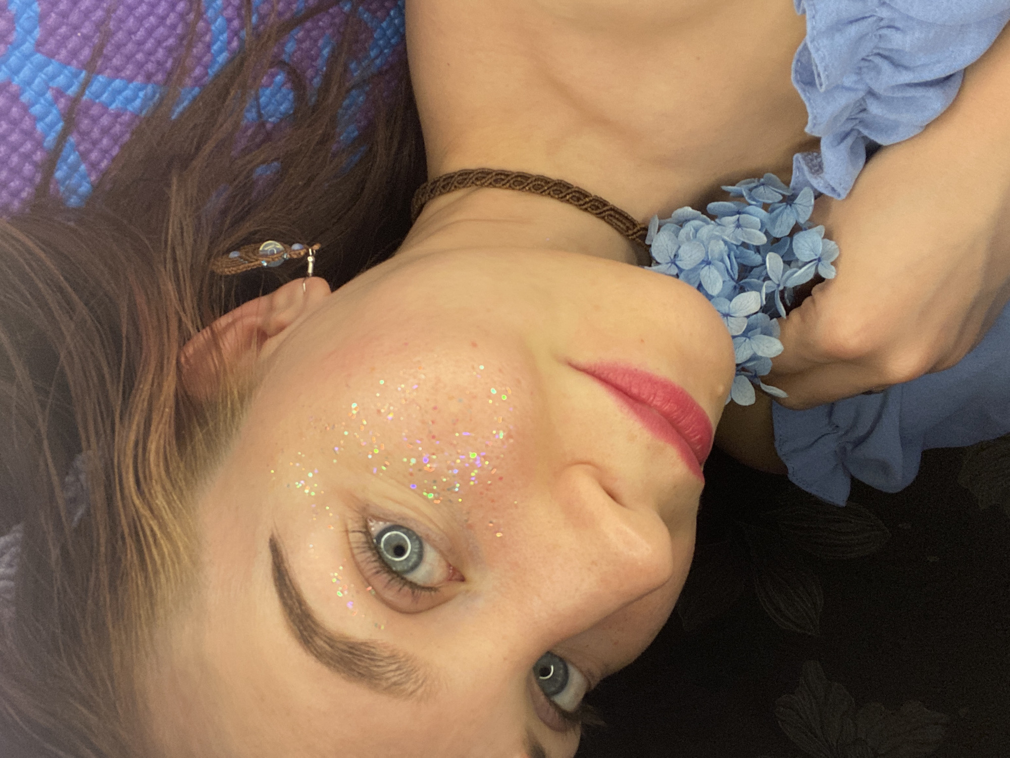 A medium close-up shot depicts a light-skinned woman with fair hair and blue eyes. Her face is adorned with glitter. She wears a brown woven choker necklace and has blue flower accents near her chest. She is also wearing an earring on the left side. Her hair is a mix of brown and blonde tones. She appears to be lying down on a surface with a purple and blue patterned section visible in the background and a floral patterned dark section. There is no specific evidence in the image to determine the exact location in the city of Chaikovsky, Russia.