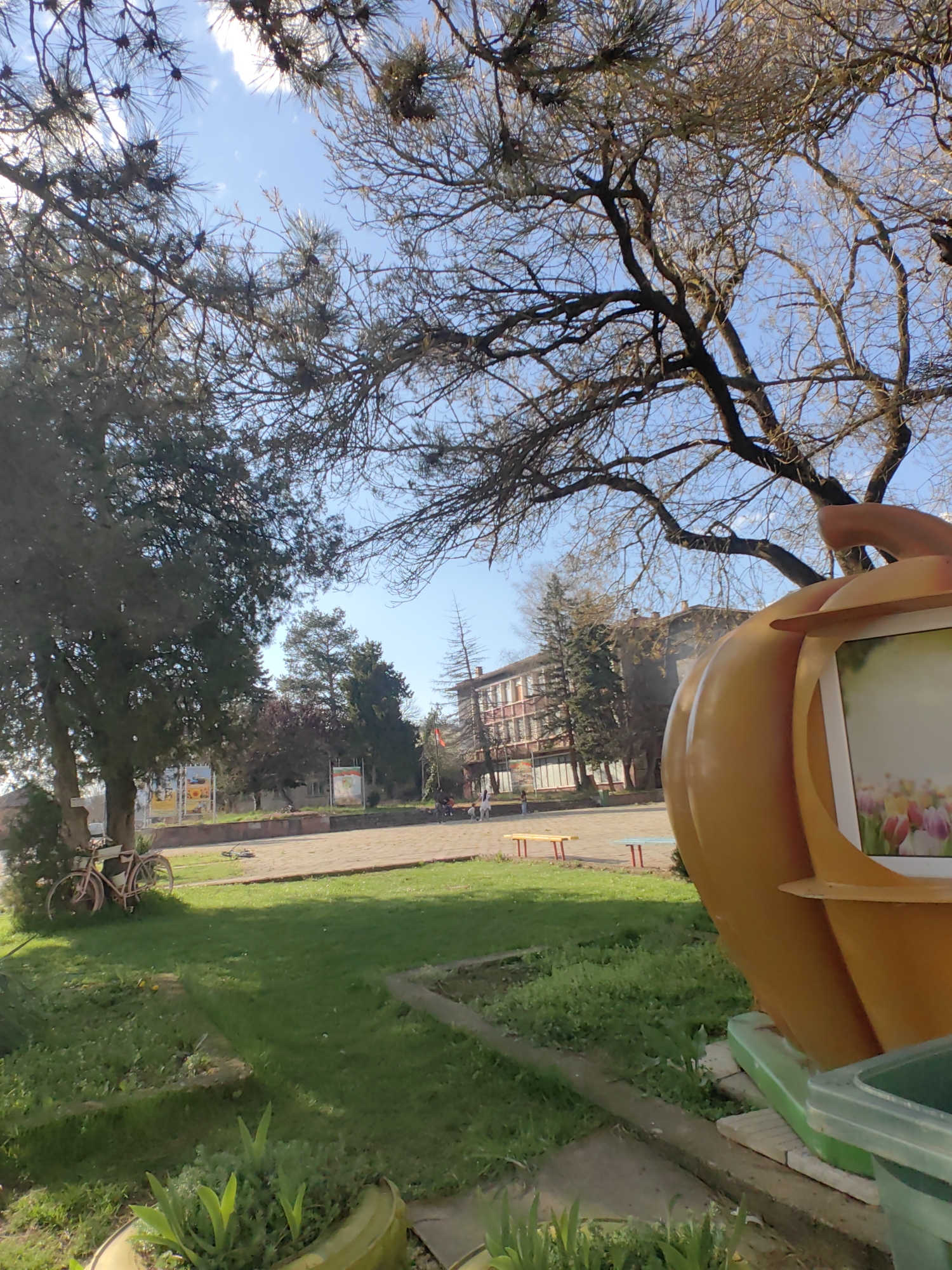 The image shows an outdoor scene in Sennik, Bulgaria, with a building in the background and trees framing the shot. A large, orange pumpkin-shaped structure stands in the foreground to the right. A bicycle is parked near a tree on the left side. The ground is covered with grass and a concrete path. People are visible in the background, walking near benches. The sky is blue with sparse clouds.