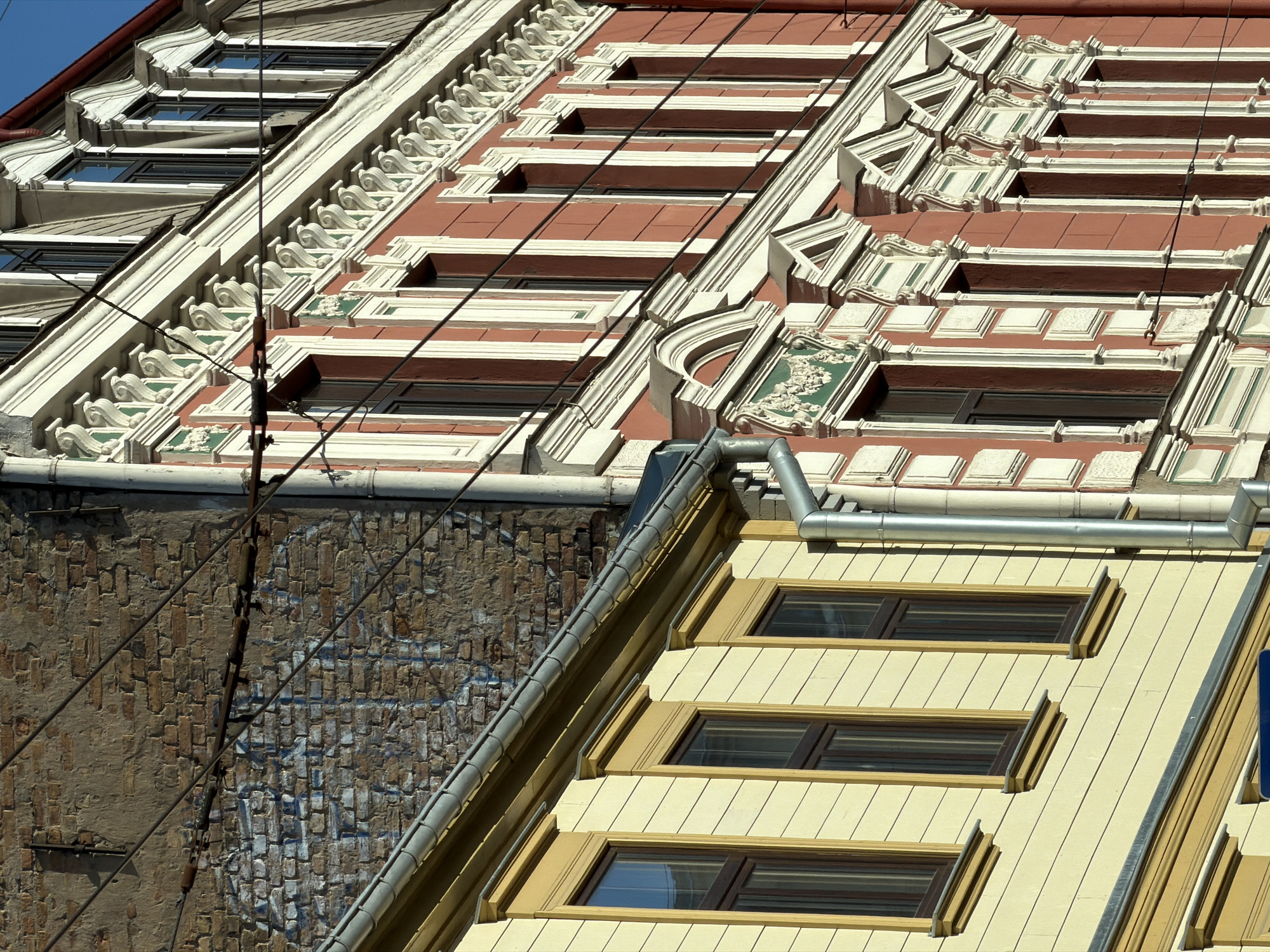 The image shows a close-up of several buildings in Riga, Latvia. The architecture is characterized by intricate facade designs, showcasing a mix of styles. The buildings feature decorative window frames and cornices, with colors ranging from red and white to yellow and brown. Some of the buildings have been updated with modern details, such as updated windows and siding. Overhead power lines are visible in the foreground. A brick wall with some graffiti-like markings is also present.