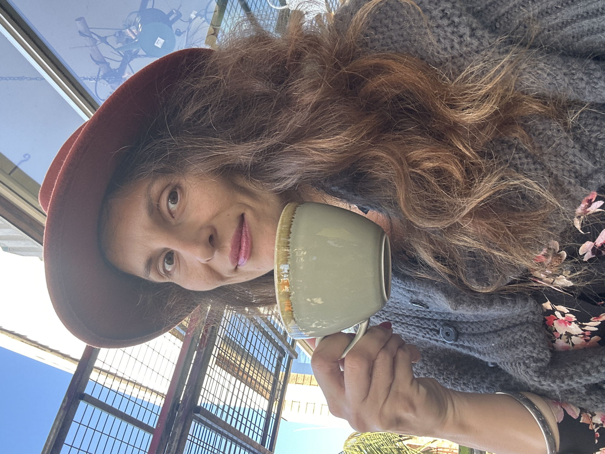 A woman, wearing a rust-colored hat, a gray knitted cardigan over a black floral dress, and a silver bracelet on her left wrist, holds a light green mug with a gold rim in her right hand to her lips. She is outdoors, with a clear blue sky overhead and a lattice-like structure behind her, suggesting an outdoor seating area. The background includes a large window reflecting the sky and some industrial elements. To the left, palm fronds are visible. The woman has long, wavy brown hair and is looking directly at the camera with a slight smile.