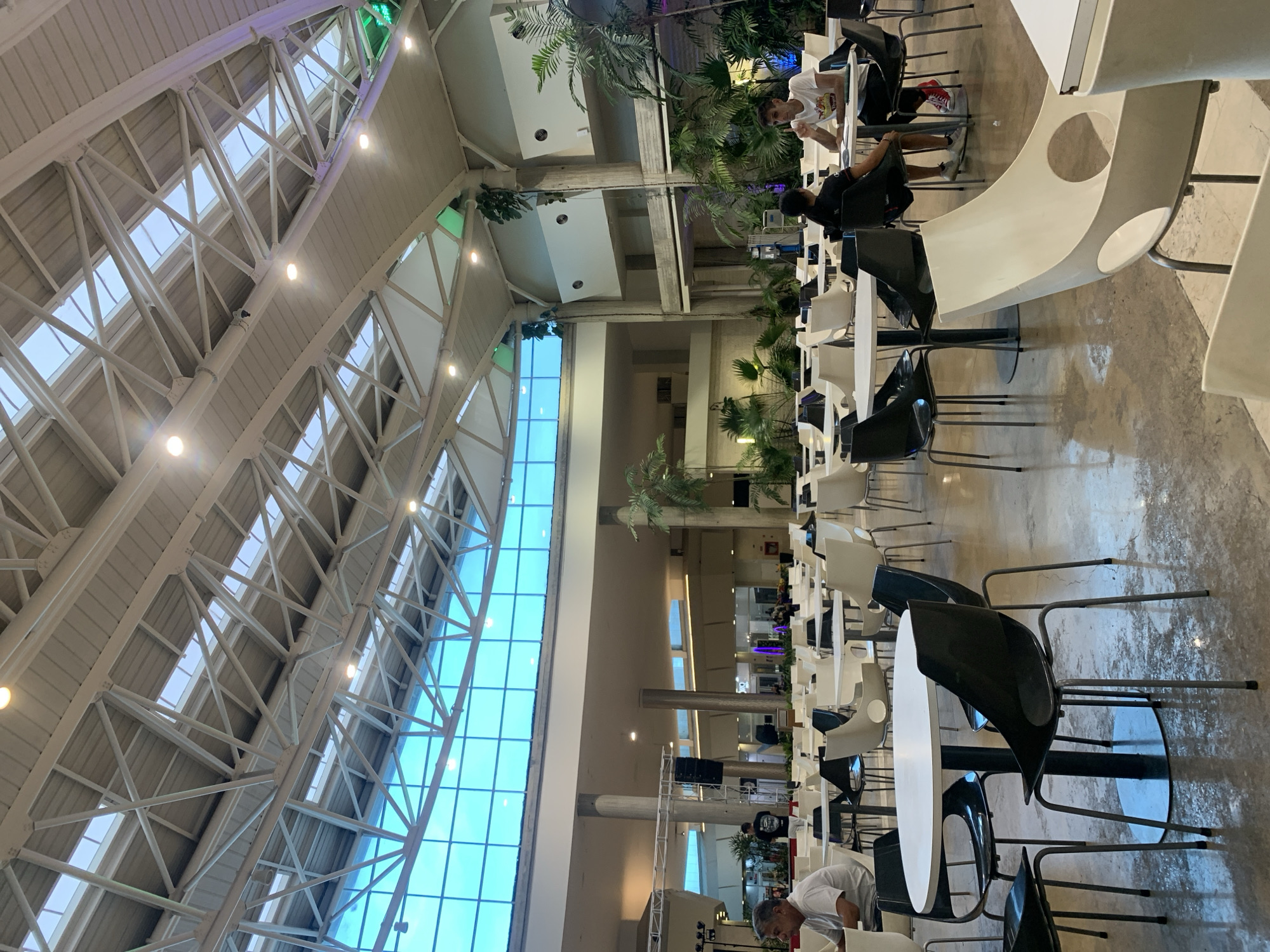 This image captures a vast, brightly lit indoor space, likely a food court or a large common area within a commercial building, located in Pampatar, Venezuela. The high, vaulted ceiling is a dominant feature, constructed with an intricate white metal truss system that supports numerous rectangular skylights, allowing ample natural light into the area. Recessed lights are also integrated into the ceiling structure.
Along one side of the building, a striking floor-to-ceiling blue-tinted glass wall or window stretches upwards, providing a vibrant color contrast to the neutral tones of the interior architecture. The space is multi-level, with balconies and upper floors visible in the background, supported by substantial cylindrical concrete columns.
The ground floor is furnished with numerous tables and chairs, primarily in black and white, featuring modern designs. Tables are typically round or rectangular with white surfaces, complemented by contemporary black or white seating. Several individuals are present, seated at the tables, appearing to be engaged in eating, socializing, or using electronic devices. Lush green palm plants are strategically placed throughout the area, enhancing the aesthetic and creating natural divisions within the large space.
The flooring appears to be polished concrete. The natural illumination from the skylights and large window indicates it is daytime. The scene conveys a spacious and relaxed atmosphere typical of a modern public gathering place. No discernible text is visible within the image.