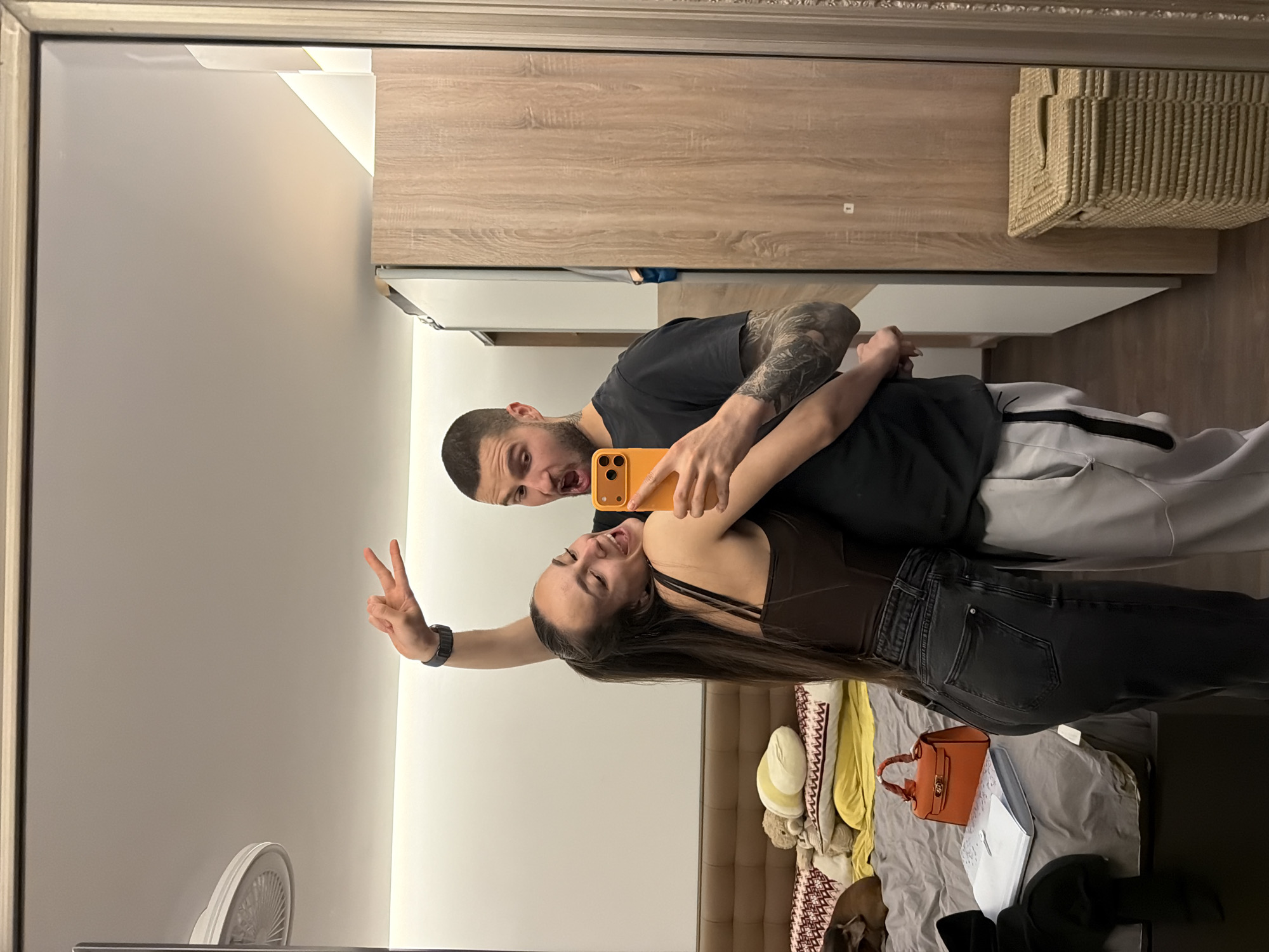 A couple is taking a selfie in a mirror. The man has a shaved head and is wearing a dark t-shirt, showing a tattoo on his arm. He is holding an orange smartphone. The woman is in front of him, wearing a brown top and dark jeans, and holding up a peace sign with her left hand. The background shows part of a bedroom with a bed, pillows, a purse, and some clothes scattered on it. Also visible in the reflection are the upper portion of a wardrobe or closet and a wall-mounted fan. There is no discernible information that places this scene specifically in the city of Sofia, Bulgaria.
