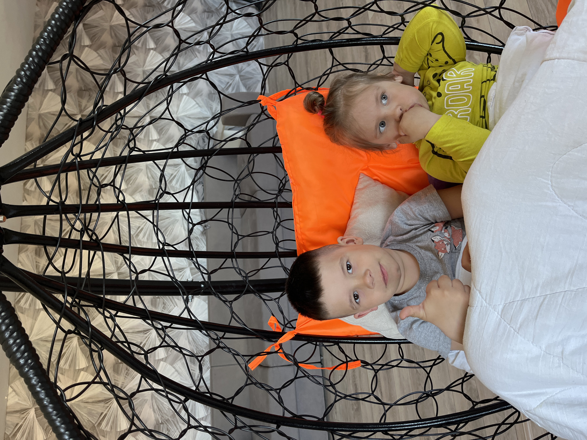 Two children are lying in a black, egg-shaped, hanging wicker chair. A girl with blonde hair is on top, wearing a yellow shirt with the word "ROAR" on it, and she is sucking on her fingers. A boy with short dark hair is below her, wearing a gray shirt with a fox graphic. They are both covered with a white blanket and lying on an orange pillow. The background shows a floor and wallpaper with a geometric pattern. The location is Chaikovsky, Russia.
