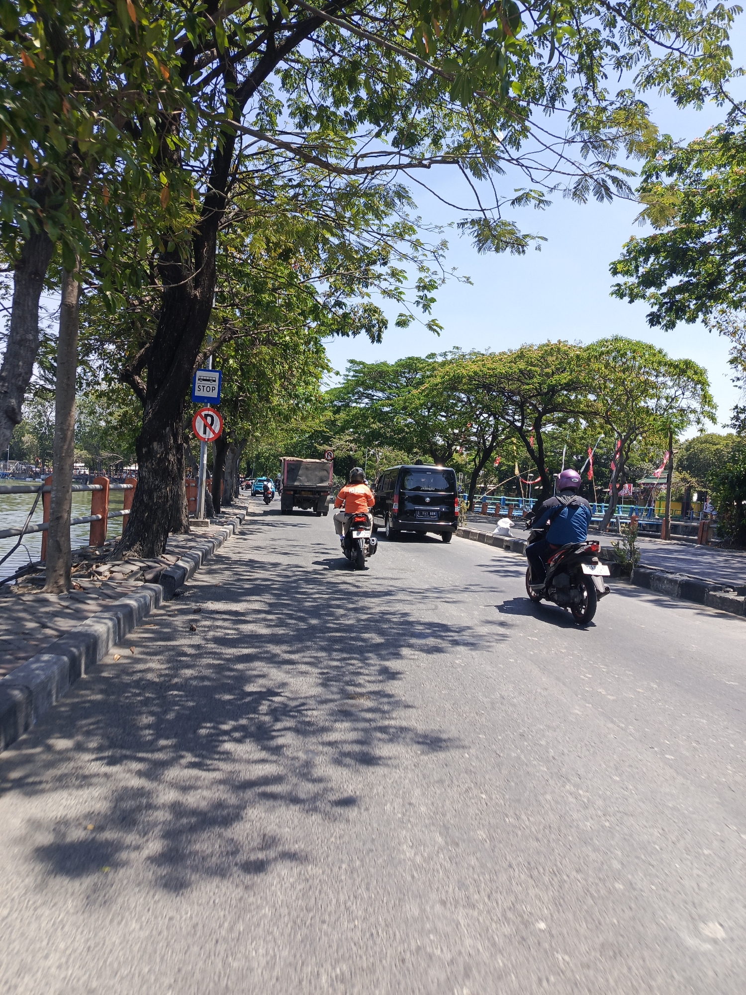 The image captures a street scene in Surabaya, Indonesia, alongside a body of water, likely a river or lake, on a sunny day. The road is paved and appears to be a main thoroughfare. Several vehicles are present, including a red dump truck, a black van, and two motorcycles. One motorcycle in the foreground is ridden by a person wearing a blue jacket and a purple helmet. Another motorcycle further back is ridden by a person wearing an orange shirt. The street is lined with mature trees that cast dappled shadows on the asphalt. On the left side of the road, there is a sidewalk with a concrete curb and a railing along the water. Traffic signs are visible, including a "STOP" sign and a "no parking" sign. In the background, more trees and some buildings can be seen, as well as decorative red and white streamers strung across the road. The overall impression is of a bustling urban environment with natural elements.