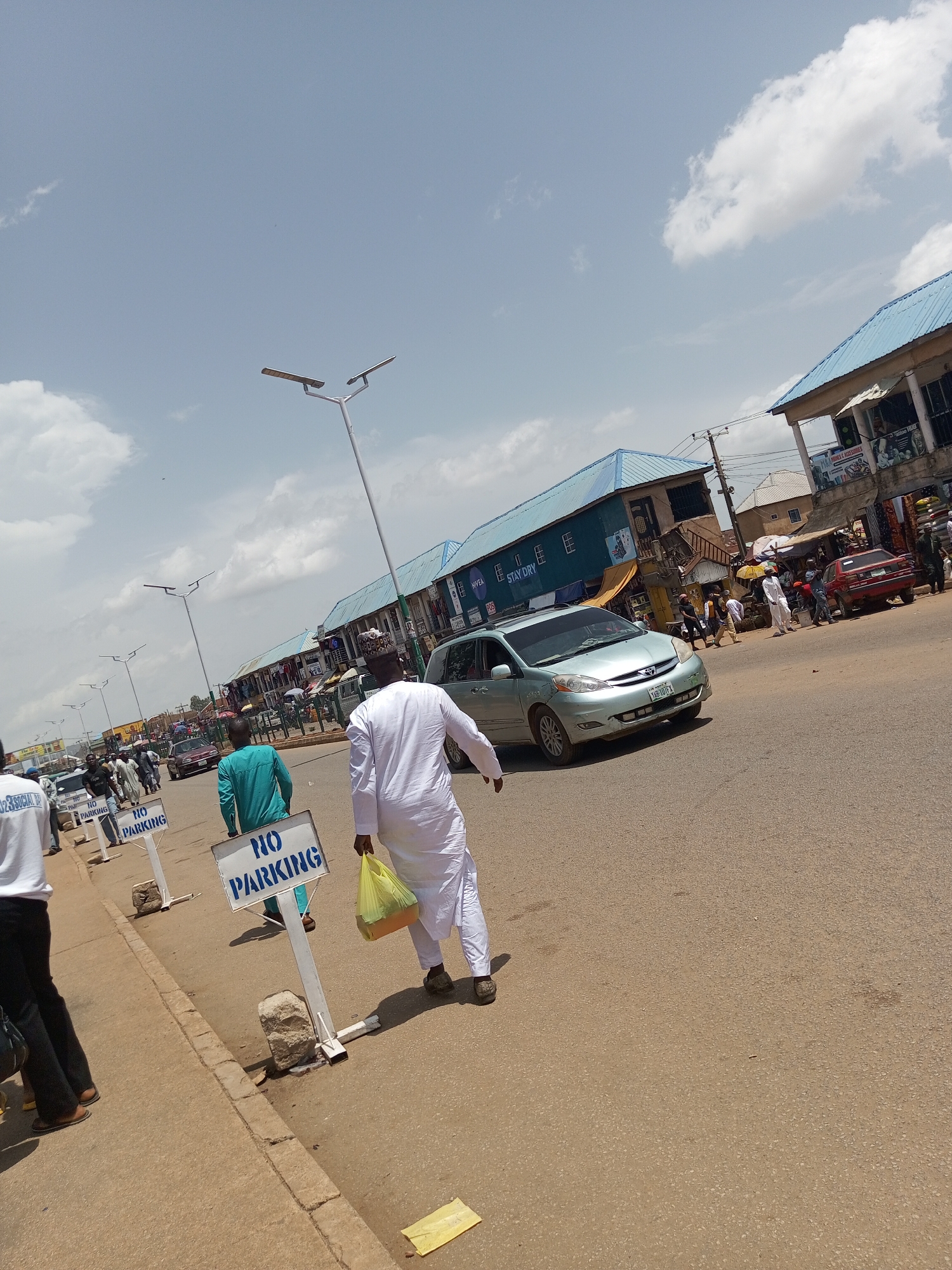 The image depicts a street scene in Bukuru, Nigeria. In the foreground, a man in white traditional attire walks away from the camera, carrying a yellow bag. To his left, another man in a teal-colored outfit is also walking, facing away. Several "NO PARKING" signs are visible on the sidewalk. The street is lined with buildings with blue roofs, some of which appear to be shops. A light blue minivan is driving on the road, and other vehicles and pedestrians are present in the background. The sky is partly cloudy.