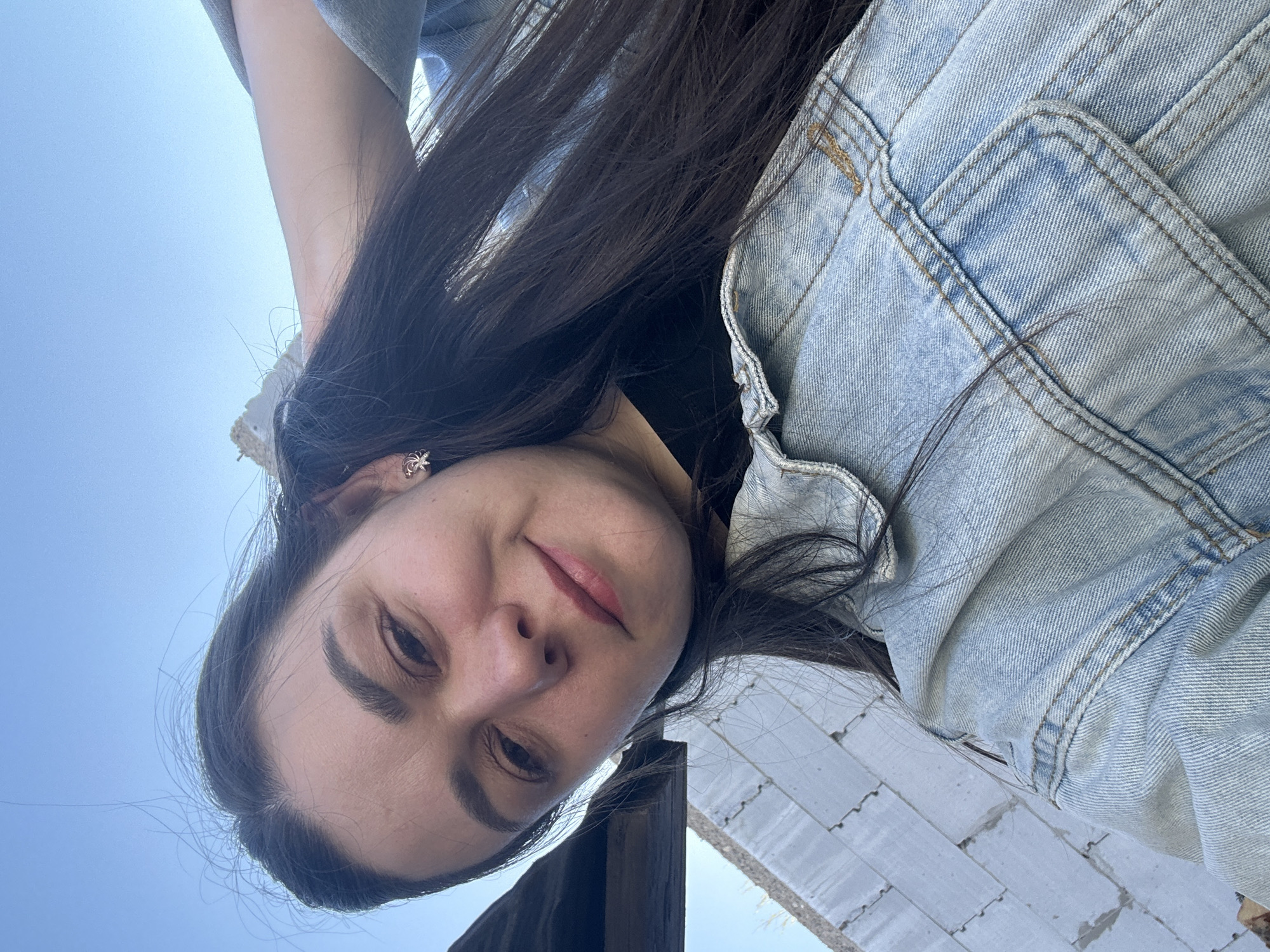 A selfie photo taken in Chaikovsky, Russia shows a woman with dark hair and a denim jacket. Her face is close to the camera, and part of a white brick structure is visible behind her. The background includes a clear blue sky.