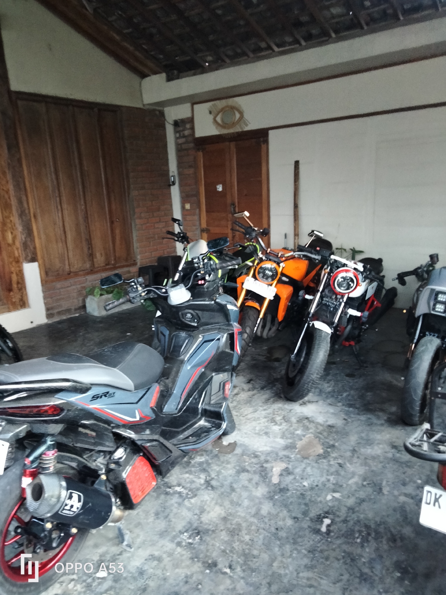 The image depicts a collection of motorcycles parked in what appears to be a covered outdoor area or garage, possibly at a residence in North Kuta, Indonesia. In the foreground, a dark-colored scooter-style motorcycle is visible, featuring a prominent black exhaust pipe with a white logo. To its right, two other motorcycles are parked side-by-side. The first is an orange motorcycle with a round headlight, and the second is a white and black motorcycle, also with a round headlight. Further to the right, another motorcycle is partially visible, distinguished by a license plate that reads "DK". The ground is a concrete surface, and the area is framed by brick walls and wooden paneling on the left. The ceiling is constructed of wooden beams and what appears to be tiles. There is a decorative circular wall hanging resembling an eye above a wooden door in the background. The lighting suggests an overcast day or shaded area. No people are present in the image.