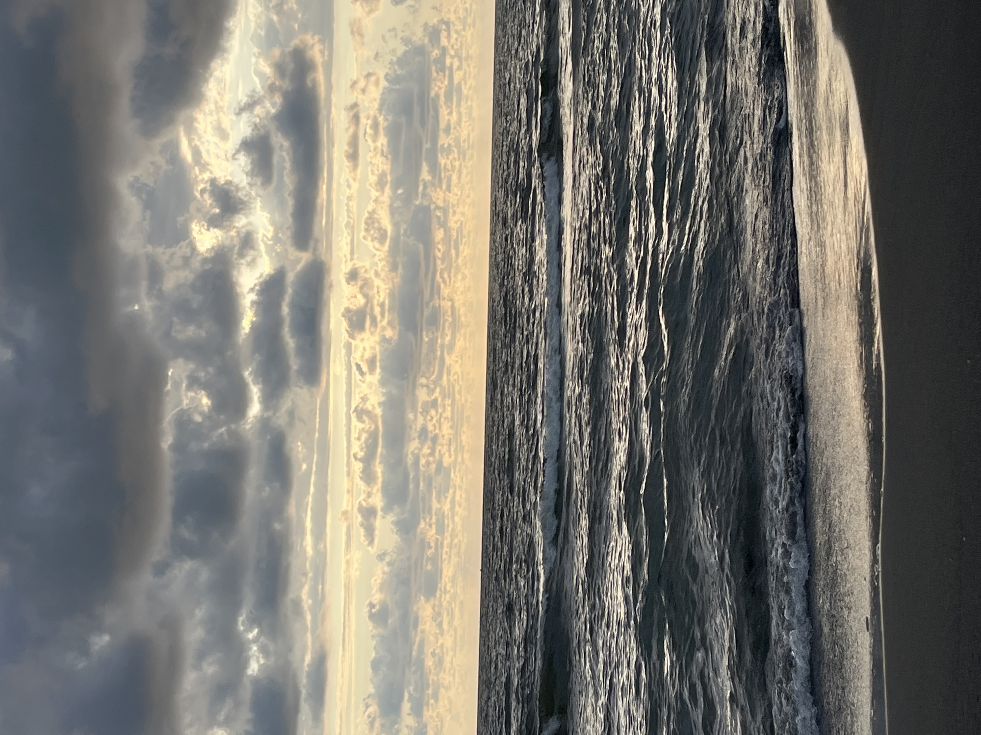 The image depicts a view of the Black Sea from the shore in the Sapsi community, Georgia. The sky is filled with dramatic, dark clouds interspersed with patches of lighter sky, suggesting either an approaching storm or a transition in weather. Sunlight peeks through the clouds, casting a soft glow on the horizon. The sea is choppy, with waves rolling towards the shore and creating white foam as they break. The foreground consists of a dark, sandy beach. No people or man-made structures are visible in the frame.