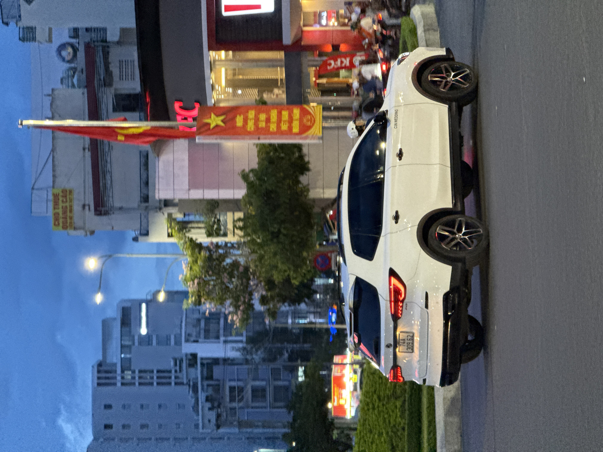 The image depicts a white SUV driving on a street in Nha Trang, Vietnam, at dusk. The sky is a deep blue with hints of clouds, suggesting the transition from day to night. Buildings of varying heights, some illuminated from within, line the street. A prominent KFC restaurant with its iconic red and white signage is visible on the right side of the image, with a Vietnamese flag displayed on a tall pole in front of it. The street is paved, and a green, manicured hedge runs along the side. A few pedestrians and motorcyclists are present in the background, moving along the street. The SUV has its headlights on, and its license plate is partially visible, showing characters and numbers. The overall scene captures a moment of urban life in Vietnam during the evening hours.