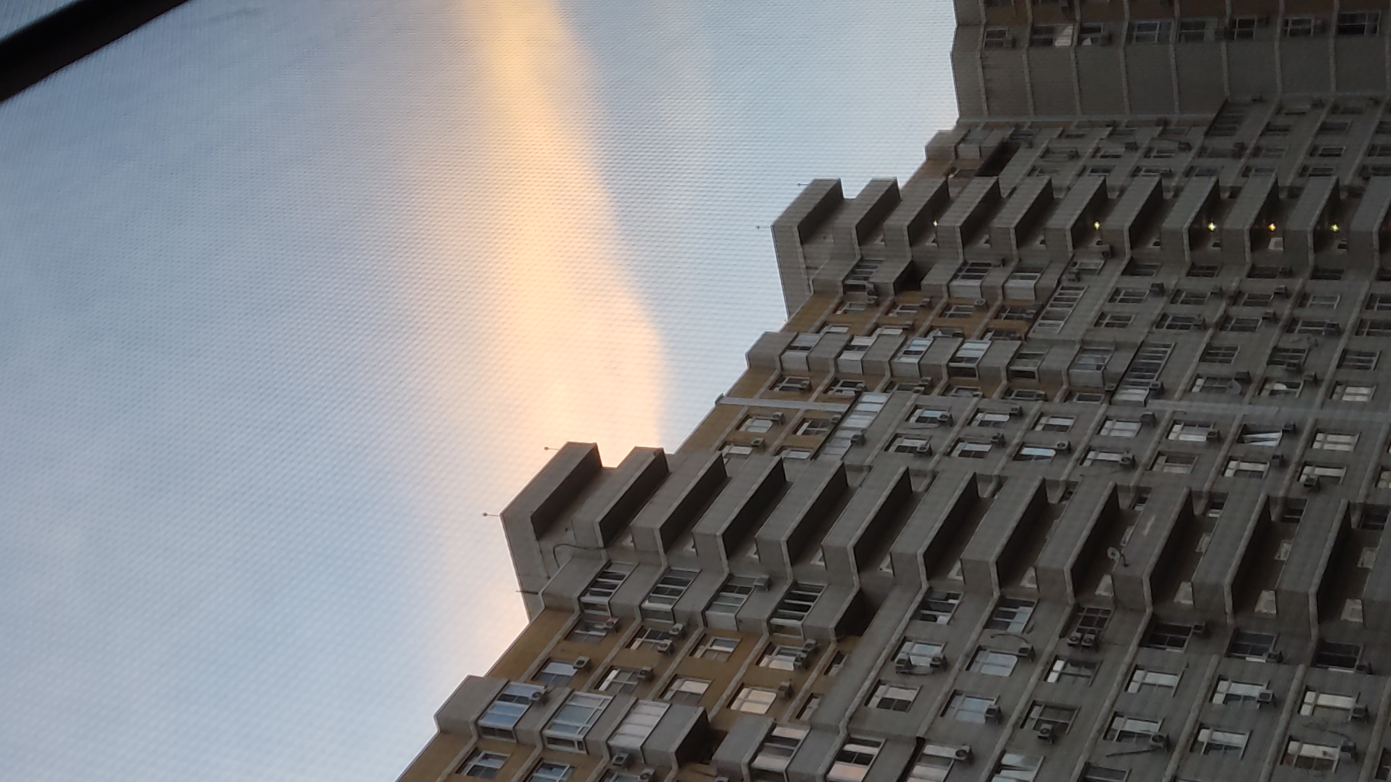 The media file displays a low-angle shot of a towering, Brutalist-style apartment building against a sky with soft, diffused light. The building's facade is characterized by repeating geometric patterns of balconies and recessed windows, creating a sense of depth and shadow. The concrete structure appears weathered, with visible air conditioning units mounted outside many of the windows, suggesting a residential setting.
The sky exhibits a gradient of pale blue transitioning to a warm, hazy orange or yellow, indicative of either sunrise or sunset. The lighting suggests a calm and possibly cool atmosphere. The scene is oriented such that the building occupies the right side of the frame, appearing to recede upwards and to the right, while the sky dominates the left. The texture of the sky, with its subtle crisscross pattern, suggests it may have been captured through a window or a screen.
No people are visible. The building itself, with its repetitive and somewhat stark design, could evoke a sense of anonymity or urban density. The location is identified as Krasnodar, Russia. There is no visible text in the image. The overall impression is one of urban architecture captured during a transitional time of day.