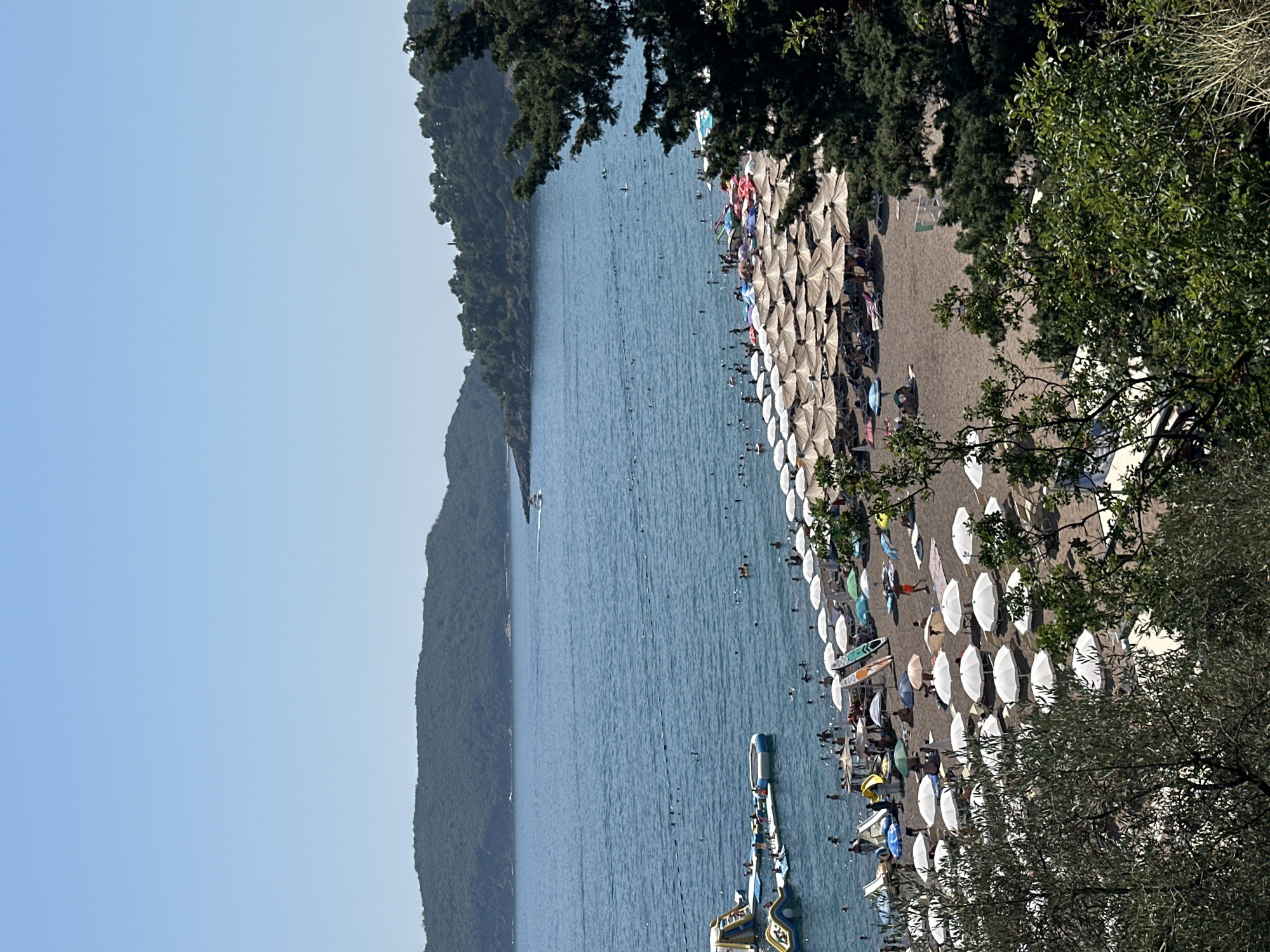 This is an aerial, eye-level, full shot of a crowded beach in Boreti, Montenegro, under a clear blue sky. The beach is filled with a vast expanse of tightly packed, beige and white umbrellas, shading numerous sunbathers on the dark sand. In the water, many people are swimming, and a few are interacting with a large inflatable water park. A speed boat is visible in the distance, and lush green hills form the backdrop to the scene. The foreground is occupied by the branches of leafy green trees.