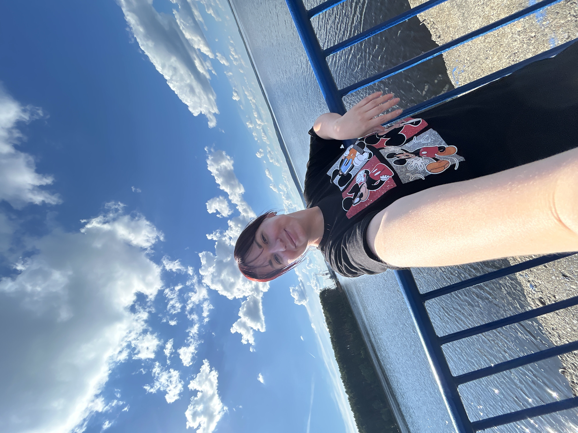 A young woman with dark hair and bangs, wearing a black t-shirt with a Mickey Mouse print, poses for a selfie on a sunny day. She is leaning on a blue railing, with a body of water behind her. The water is calm, reflecting the bright blue sky filled with fluffy white clouds. On the opposite side of the water, a dense green forest borders the shoreline, with industrial cranes visible in the distance. The location appears to be a waterfront promenade or a bridge, likely within the city of Chaikovsky, Russia, given the context. The overall atmosphere is relaxed and pleasant.