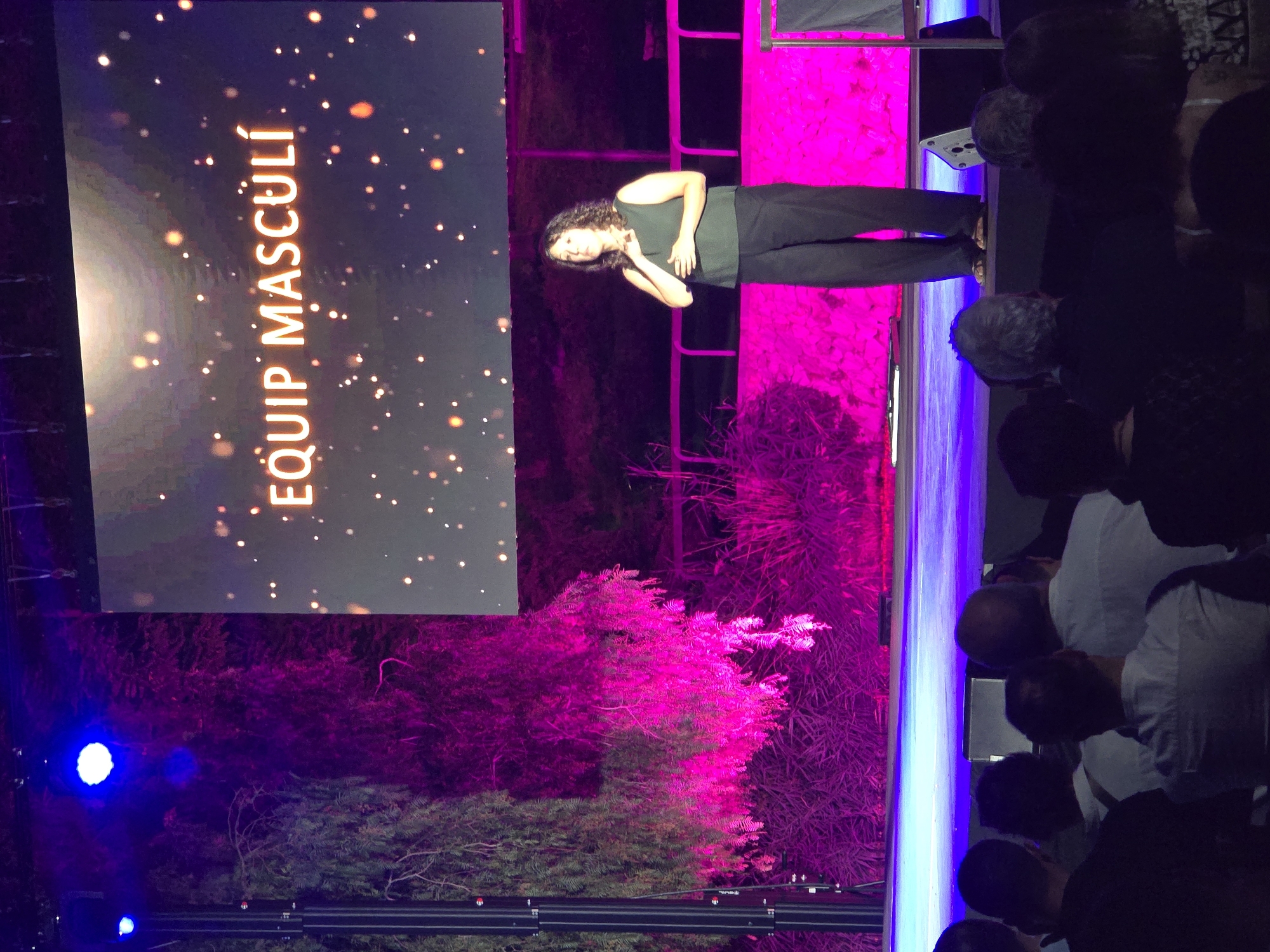 The image captures a live presentation event in Cornellà de Llobregat, Spain. A woman, likely a speaker, stands on a stage with her left hand on her chest and her right hand resting on her abdomen, facing an audience seated in darkness. She is wearing a sleeveless dark top and black pants. Behind her, a large screen displays the text "EQUIP MASCULÍ" in illuminated orange lettering against a dark background dotted with small, glowing circles, resembling bokeh effects. The stage is illuminated with vibrant pink and blue lights, casting a dramatic glow on the surrounding vegetation and the edge of the stage. The audience members are seen from behind, their silhouettes and the backs of their heads visible in the foreground. The overall atmosphere suggests a formal or semi-formal event with a focus on the speaker and the projected message.