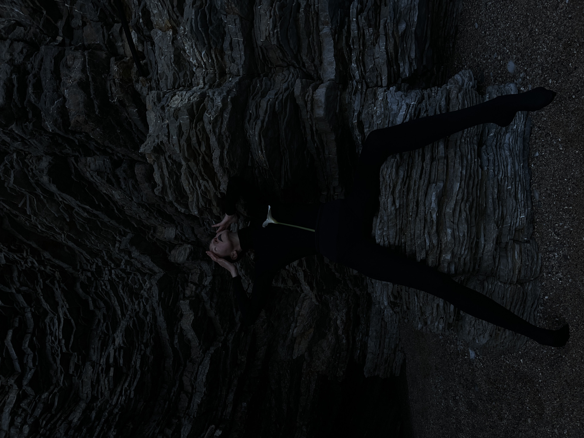A woman dressed in black tights and a long-sleeved top is lying on a rocky surface. Her body is angled across the frame, with her head positioned near the upper-left corner. Her hands are raised, framing her face. A white flower lies across her torso. The background is composed of layered, dark rock formations. The environment appears to be a cave or grotto near a beach in Budva, Montenegro.