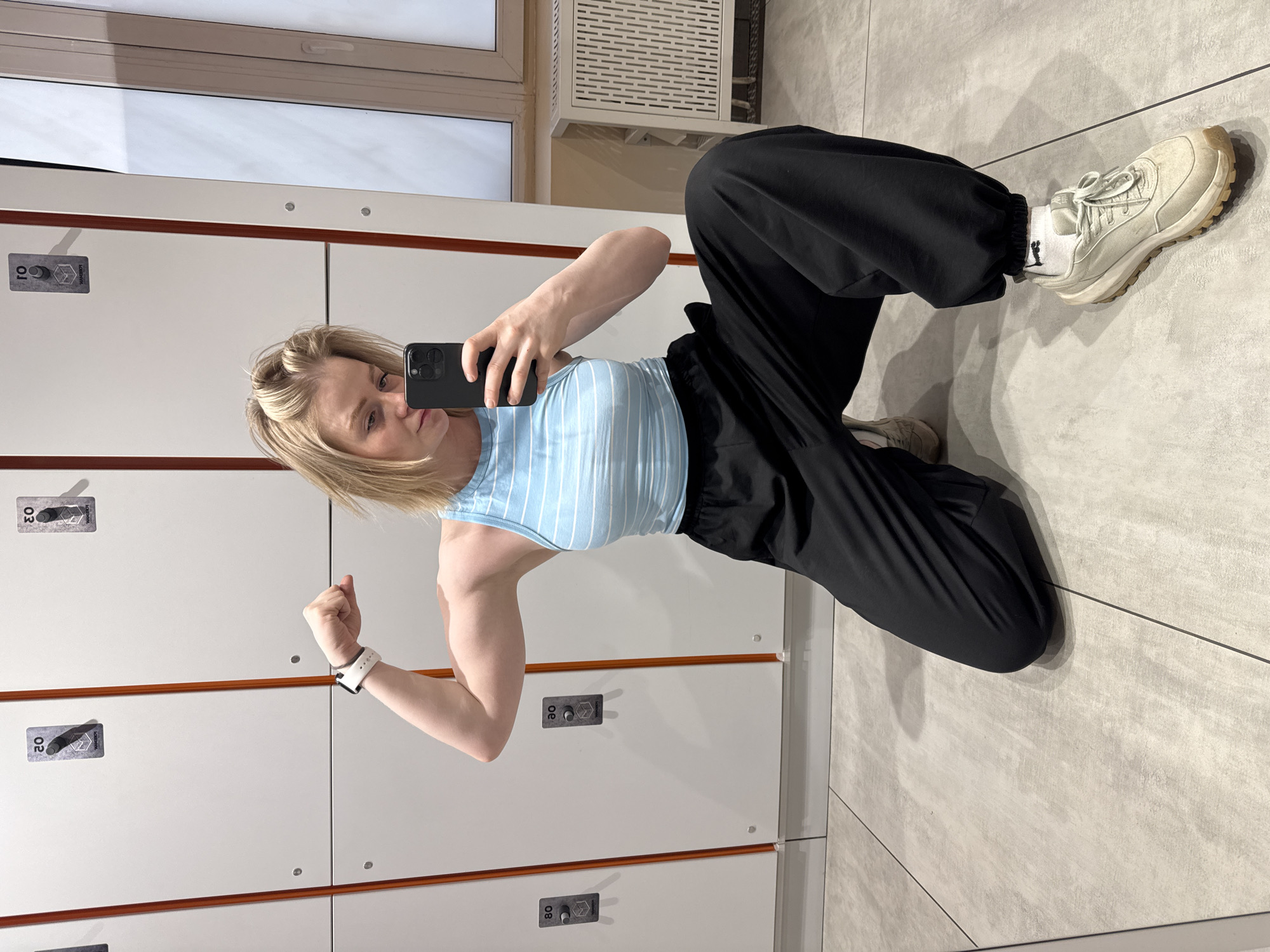 A female squats in front of a wall of lockers, inside a room with tiled floors, likely a locker room at a gym. The woman holds up her phone and takes a photo of herself with her arm flexed. She wears a blue-and-white striped tank top and loose-fitting black pants with white sneakers. The setting of the photo is not in the city of Saint Petersburg, Russia.