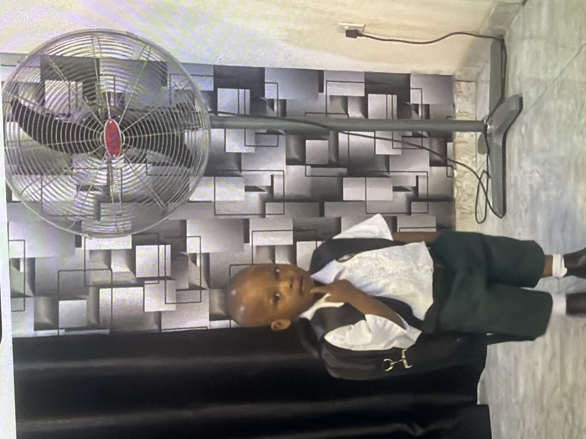 A young boy, appearing to be about 4-6 years old, stands in front of a patterned wall. He is wearing a white collared shirt with a dark vest over it, dark green shorts, white socks, and dark shoes. He has a black backpack on, and his right hand is raised to his chin in a pensive pose. To his right, a large, industrial-style fan stands on a metallic base. The fan's blades are visible through its protective grille. The background features a wall with a geometric pattern of overlapping squares and rectangles in shades of gray and white. A black curtain hangs on the left side of the image. A power cord snakes from the fan towards an electrical outlet on the right side of the image. The flooring appears to be a light-colored tiled surface. The lighting suggests an indoor environment.