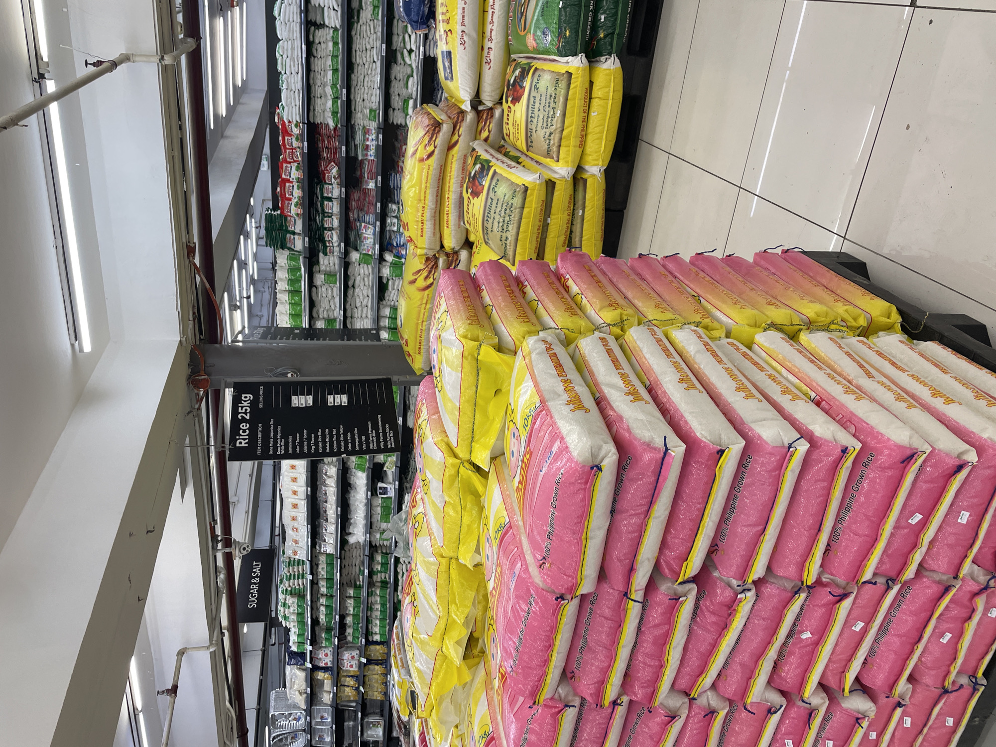 The image depicts the rice aisle of a supermarket in Davao City, Philippines. Large stacks of 25kg rice sacks are displayed on pallets, predominantly in pink and yellow packaging. The pink sacks are labeled "100% Philippine Grown Rice" and feature cartoon images. The yellow sacks have varied labels including "KOHAKU PLUS" and "Julisome Premium". In the background, shelves are stocked with more rice bags and other grocery items. A sign overhead reads "SUGAR & SALT", and another, partially visible, indicates "RICE 25KG" with a list of rice varieties. The environment appears to be a well-lit supermarket interior with tiled flooring. No people are visible in the frame.