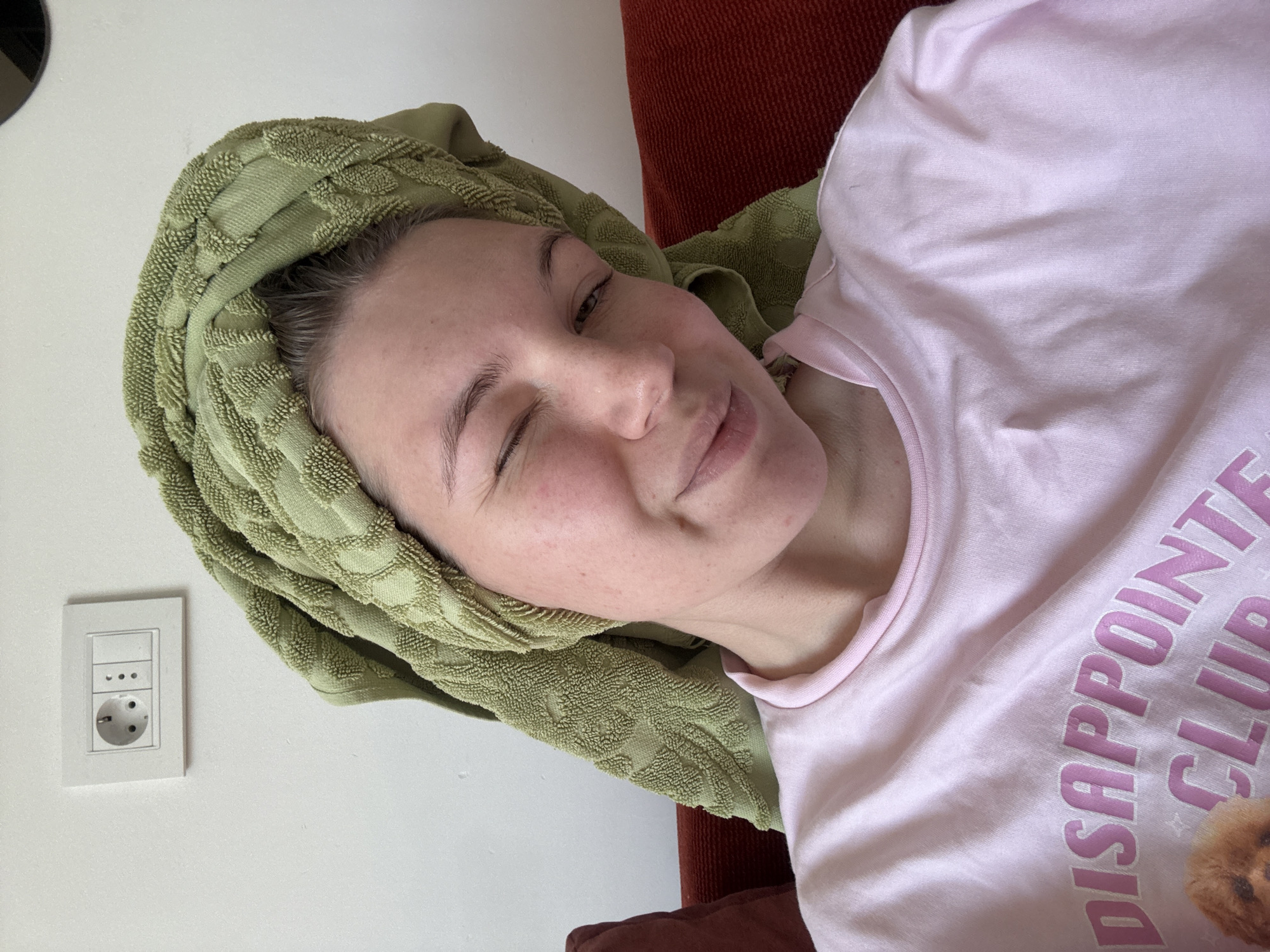 A young woman with a green towel wrapped around her head is lying on a red-brown couch. She is winking and wearing a light pink t-shirt with the text "DISAPPOINTED CLUB" printed on it. Behind her is a white wall with a light switch and European-style electrical outlet. The location appears to be indoors. The geographical location in Vozdovac Urban Municipality, Serbia, is not verifiable from the image alone.