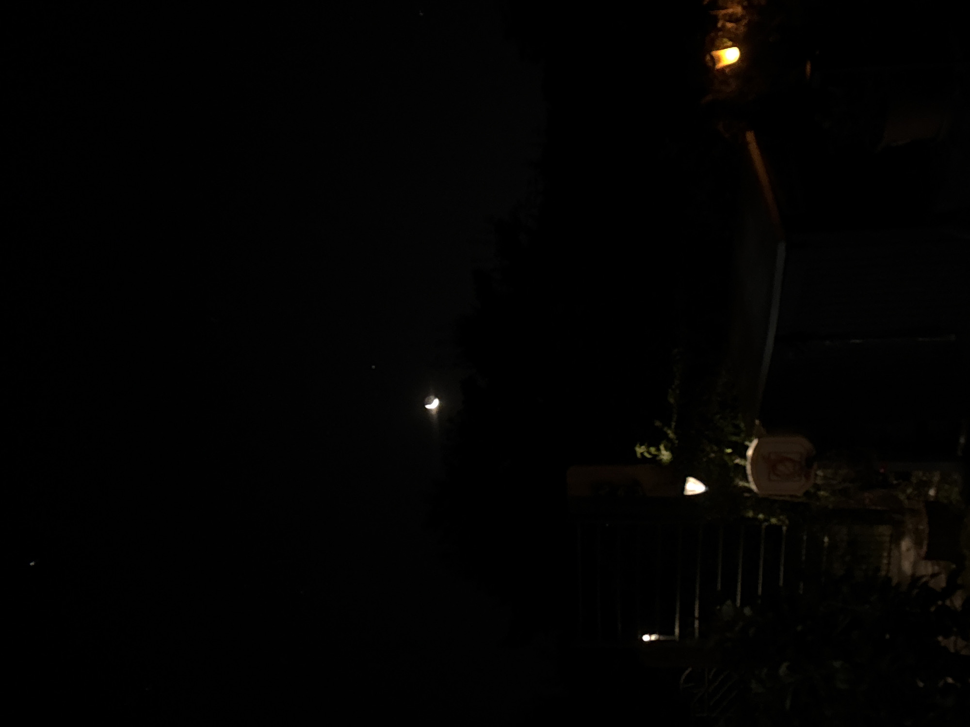 The image shows a dark night scene, likely in an outdoor area. In the upper portion of the image, a crescent moon is visible against a black sky, with a few faint stars. Below the moon, dark shapes suggest trees or distant buildings. In the lower part of the image, artificial lights illuminate parts of the foreground. There is a set of dark stairs or a railing on the left, with a light source visible. To the right of the stairs, a circular object with a red border and a light inside is present, possibly a sign or a decorative item. Further to the right, another light fixture emits a warm yellow glow, illuminating some foliage. The overall impression is of a dimly lit outdoor space at night. There are no people visible in the image. The location in the city of Lübeck, Germany, is not discernible from the visual information presented.
