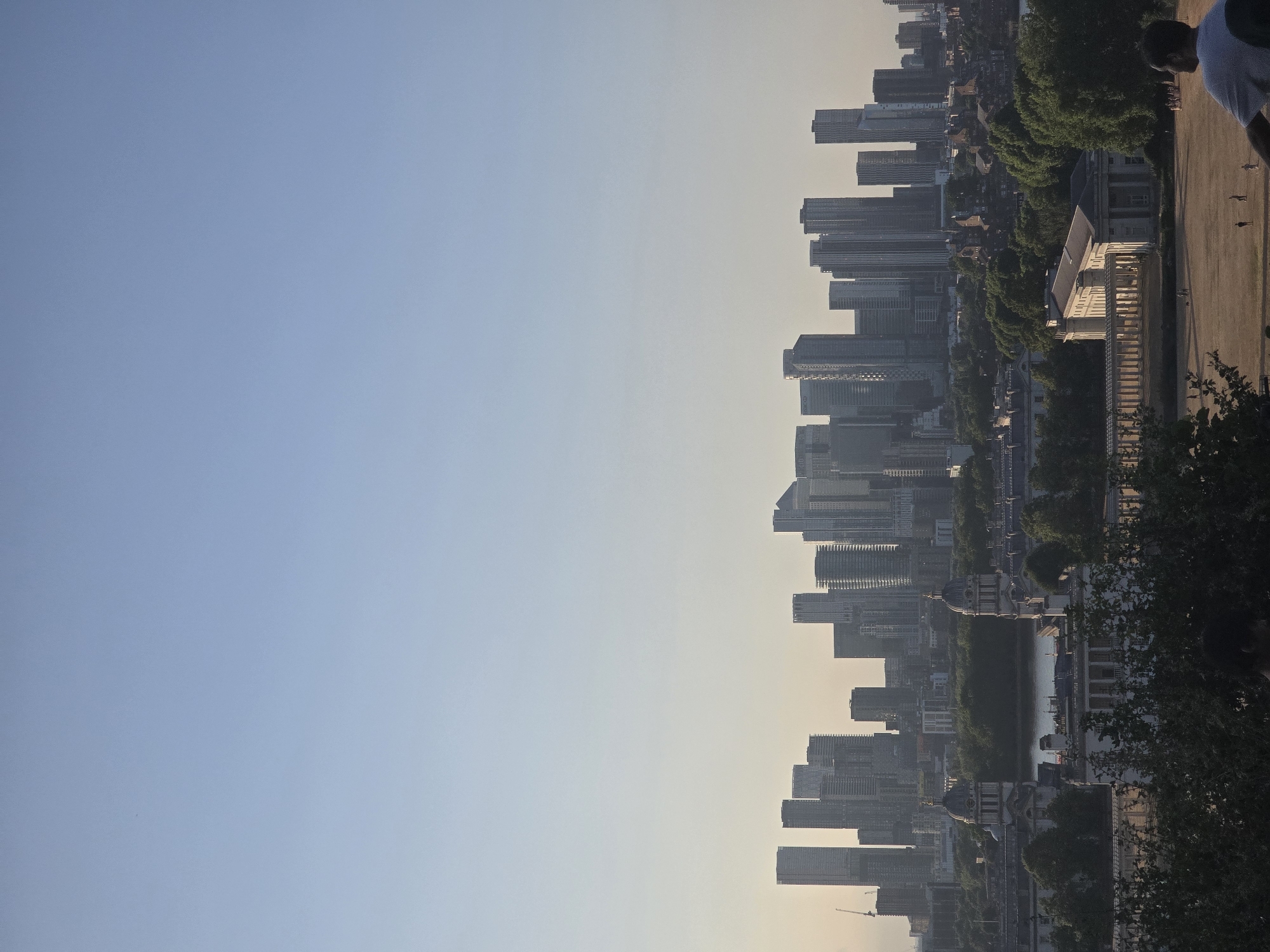The image shows a panoramic view of the Canary Wharf skyline in London, United Kingdom. In the foreground, there are lush green trees, and beyond them, a vast expanse of dry, light brown grass, with several small figures of people scattered across it, some of whom appear to be walking or engaged in activities. A notable structure in the mid-ground is a long, white building with a colonnade, and further in the distance, the River Thames flows, separating the foreground from the iconic skyscrapers of Canary Wharf. The sky above is a clear, pale blue with a gradient towards a lighter hue near the horizon, suggesting it is either early morning or late afternoon. The architecture of the buildings in Canary Wharf is characterized by modern, high-rise structures with glass facades. The overall scene captures a peaceful urban landscape with a blend of historical and contemporary elements.