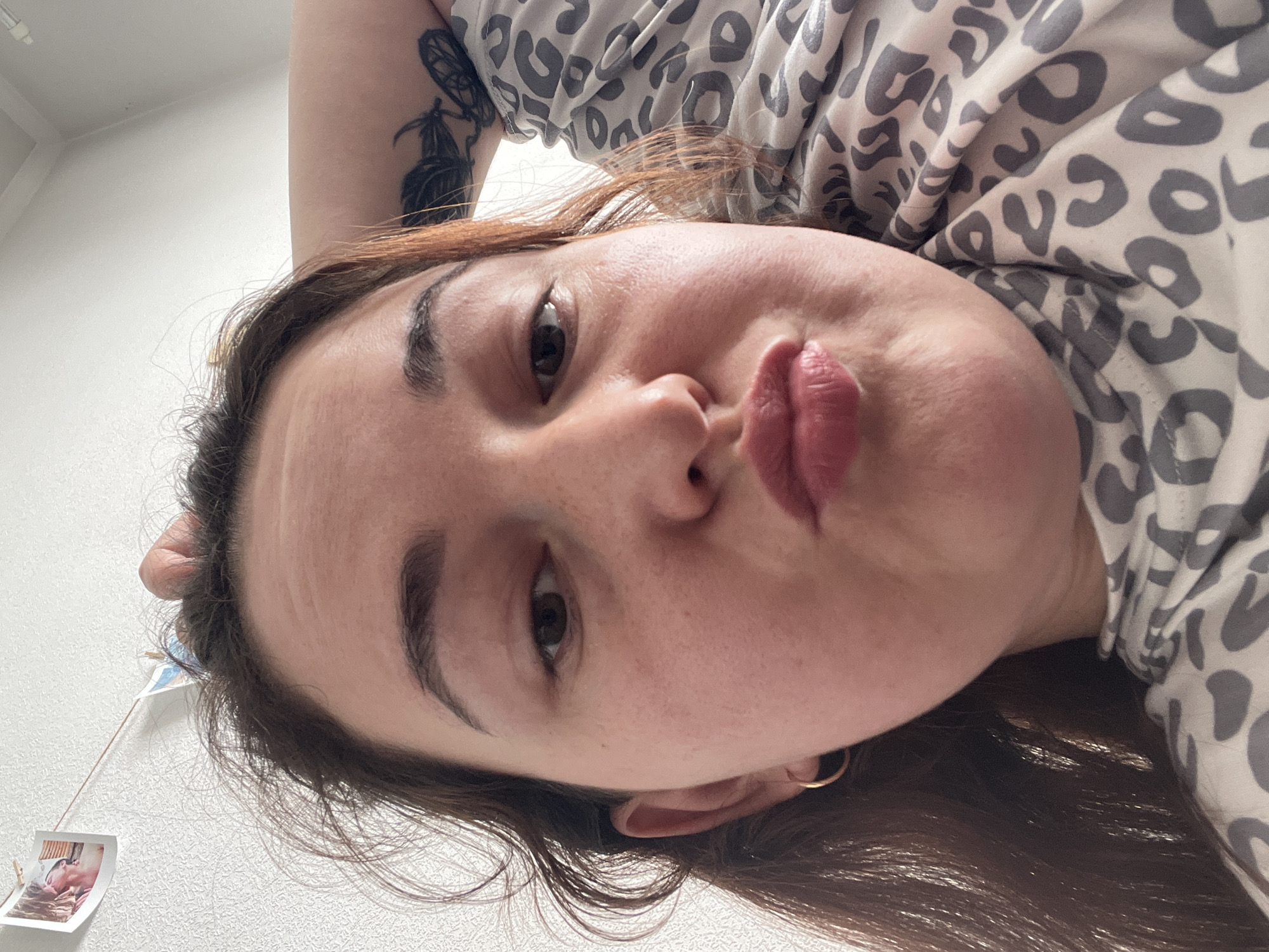 The image shows a close-up, low-angle selfie of a young adult with brown hair and fair skin. The subject is making a kissing face. They are wearing a gold hoop earring in their left ear and a grey and white leopard-print shirt. A tattoo of what appears to be a moth or butterfly with feathers is visible on their upper right arm. In the background, a white textured wall is visible, with two small photos clipped to a string hanging on the wall. The lighting suggests an indoor setting, likely a home. There is no discernible indication of the image being taken in Chelyabinsk, Russia.