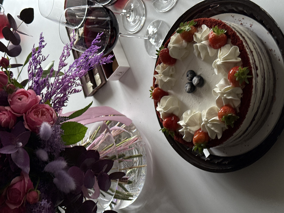 An overhead-angle shot reveals a festive tabletop arrangement. In the foreground, a round red velvet cake, approximately eight inches in diameter, is presented on a dark, possibly black, serving tray. The cake features visible red crumb layers and is generously topped with white whipped cream rosettes, fresh red strawberries, and three dark blueberries clustered in its center. To the left, a large, clear glass vase holds a vibrant floral bouquet comprising numerous pink roses, light purple feathery flowers, and dark purple foliage. A light pink ribbon is tied around the vase. To the right, two clear stemmed wine glasses, each containing a quantity of dark red liquid consistent with red wine, are visible. Behind the glasses, an open brown box displays multiple individual chocolates or pralines, and a smaller white box, possibly bearing the letters "DY," sits nearby. All items rest on a smooth, light-colored surface, likely a white tablecloth or table. The specific location within the city of Yerevan, Armenia, cannot be determined from the visual information provided in the image.