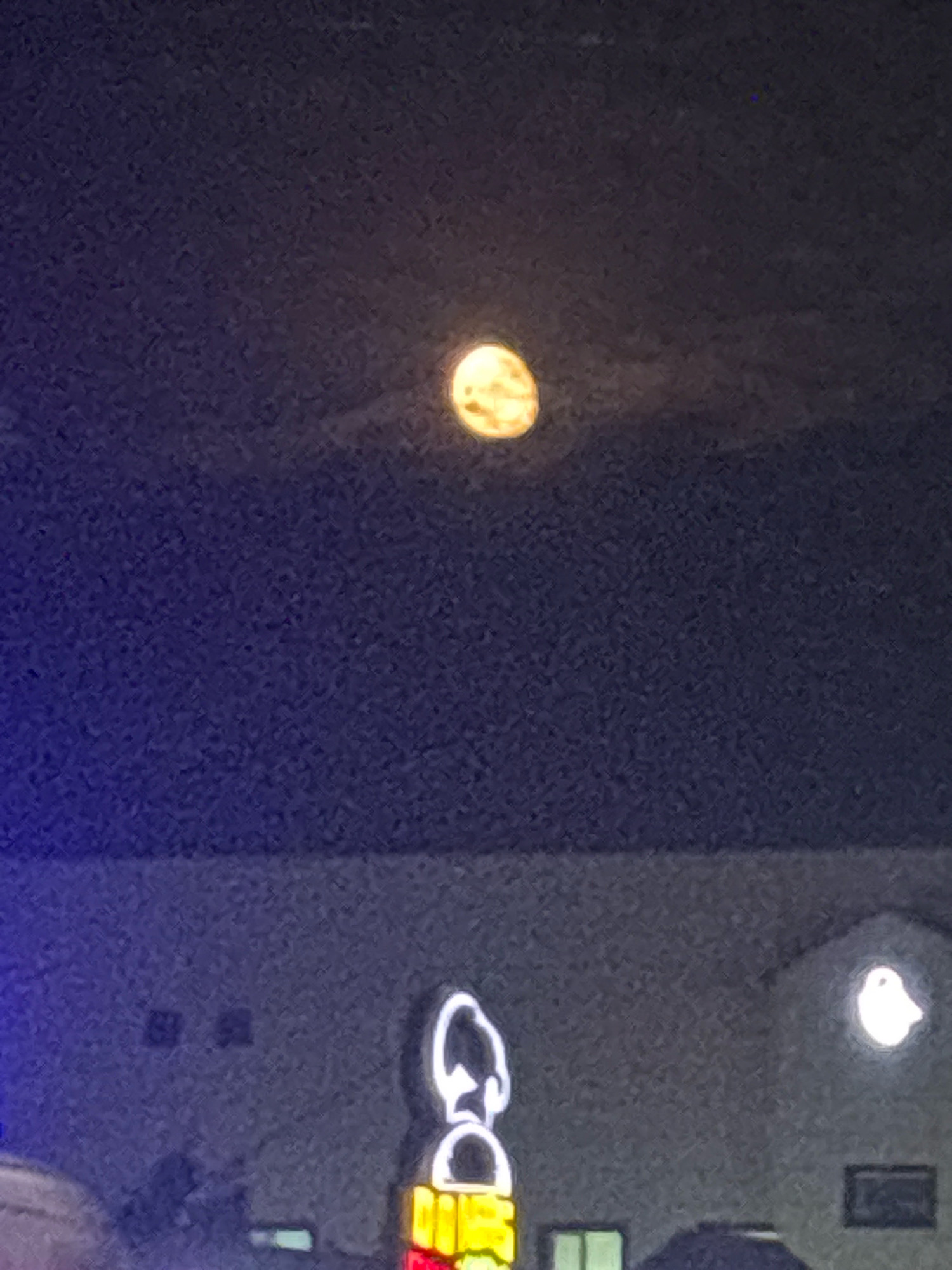 The image shows a night scene with a full moon visible in the dark sky, partially obscured by clouds. Below the moon, there is a building with illuminated signs. One prominent sign appears to be a cartoon character outline illuminated with white light, with a colorful sign at its base. Another smaller illuminated sign, shaped like a bird, is visible on the right side of the building. The building's facade is a muted gray, with a few windows and what appear to be vents or fixtures. The overall impression is that of a commercial or entertainment venue at night. Without further information, it is not possible to ascertain the specific location in the city of Taraz, Kazakhstan.