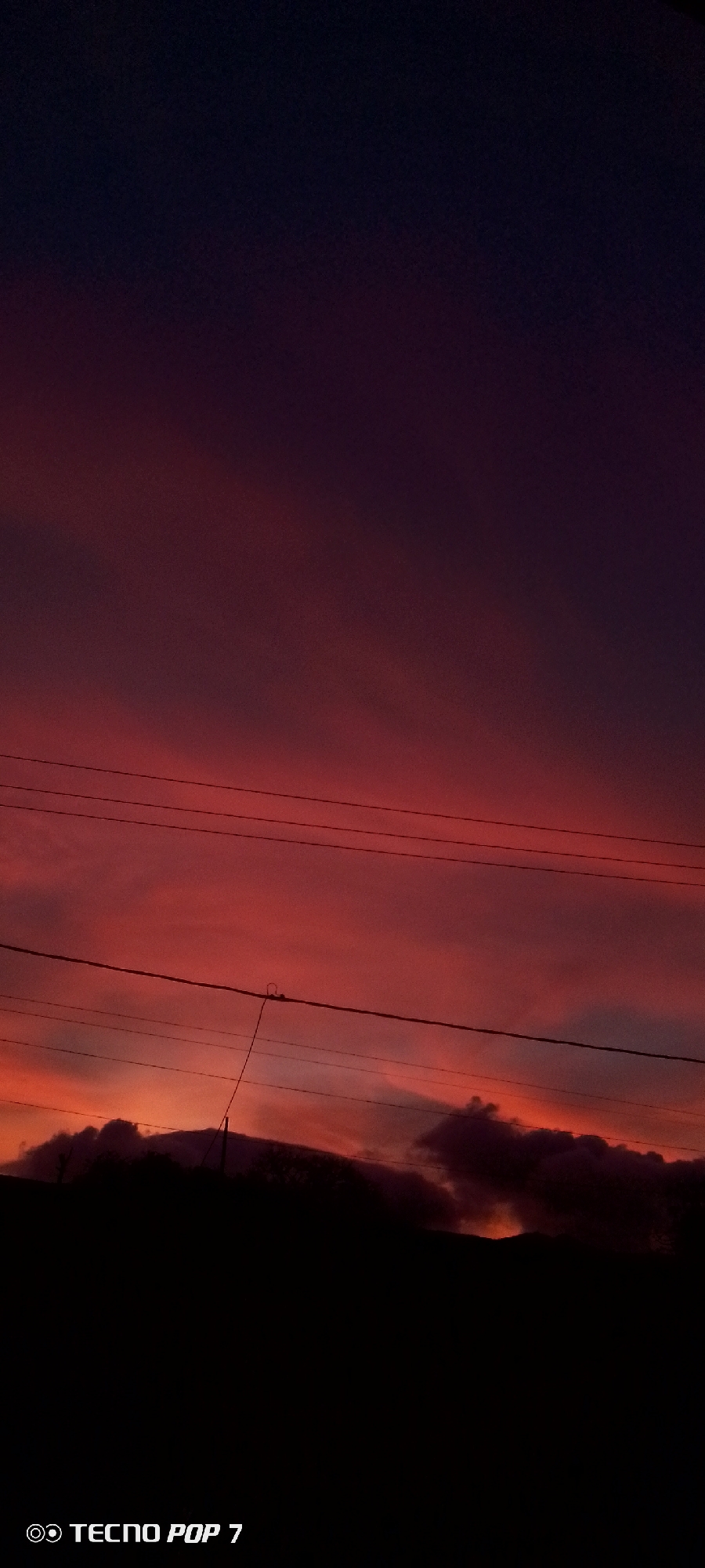 The image depicts a sunset in Altagracia, Venezuela. The sky transitions from a deep purple-blue at the top to shades of red and orange near the horizon. Several power lines stretch across the middle of the frame. Silhouetted hills or trees are visible at the bottom. The words "TECNO POP 7" appear in white at the bottom left corner of the image.