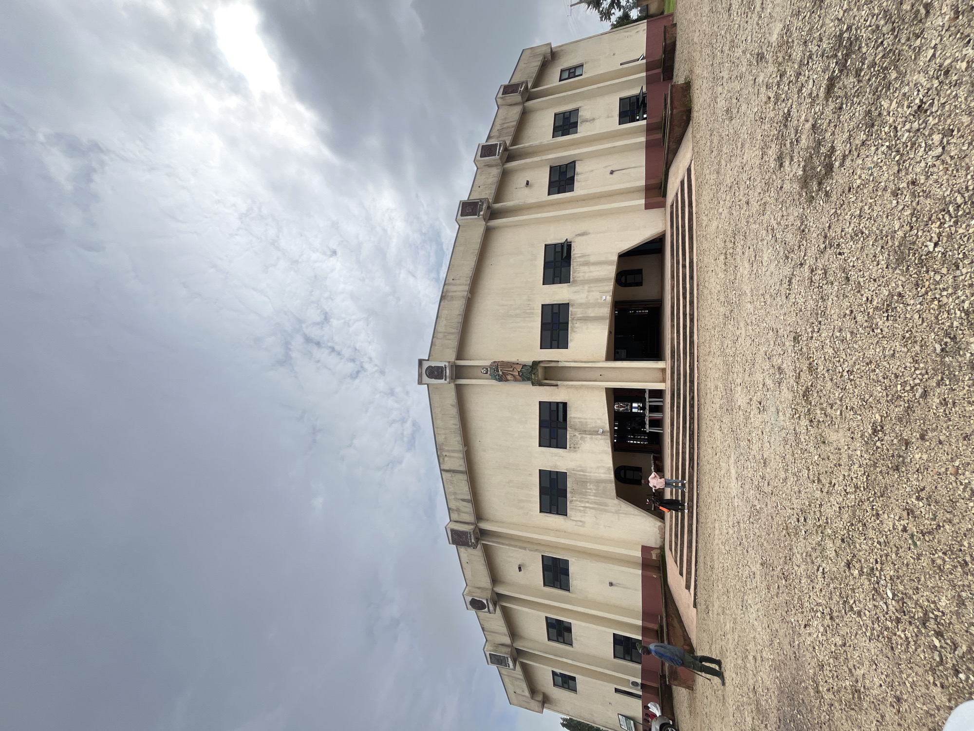 The image shows a wide-angle view of the exterior of a church building in Vom, Nigeria, under a cloudy sky. The church has a cream-colored facade with a unique architectural design, featuring angled walls and a series of dark-tinted rectangular windows. The entrance is recessed, with steps leading up to a wide opening. A statue of a saint is visible above the entrance, flanking a religious iconography.
In the foreground, the ground is covered with gravel and small stones. Two children, a girl in a pink top and a boy in a dark shirt, are standing on the steps of the church. To the left of the steps, a man wearing a blue shirt and dark trousers stands with a stick. Further to the left, a white car is partially visible. The overall scene is outdoor, with the church as the central subject.