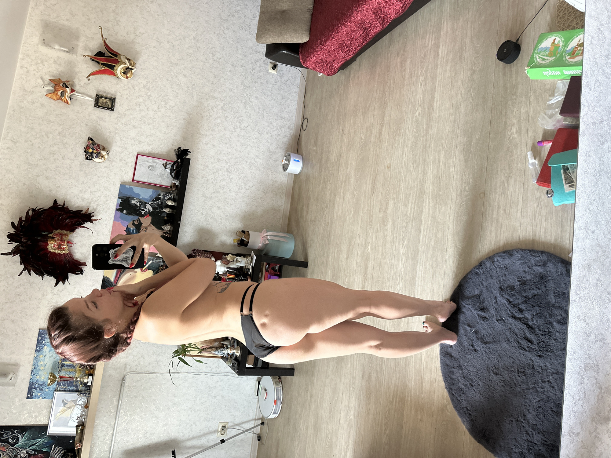 A woman is taking a mirror selfie in what appears to be a living room. She is wearing black bikini bottoms and is facing away from the camera, looking at her phone. The room has light-colored walls with decorative masks hanging on them. There is a shelf with various items on it, including artwork and trophies. A round, dark gray rug is on the floor in front of her, and a robotic vacuum cleaner is visible in the background. To the right of the image, a couch with a red textured throw is partially visible. The floor is made of light-colored laminate.