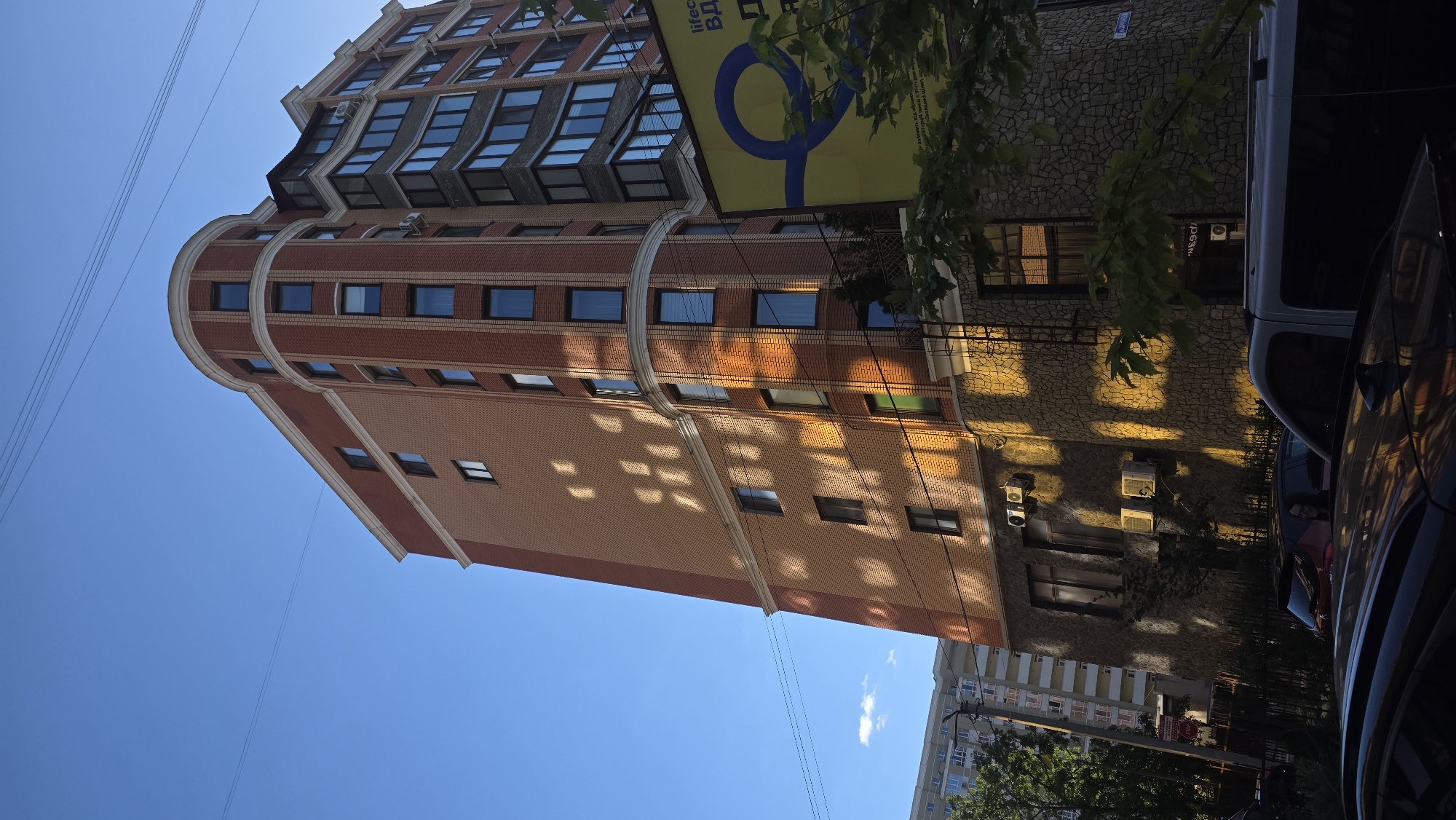 The image shows a modern, multi-story brick building in the city of Khmelnytskyi, Ukraine. The building has a curved facade and numerous windows. Sunlight casts dappled shadows on the brickwork. Power lines are visible in the sky. In the foreground, several cars are parked. A large yellow advertising banner with a blue circular logo is attached to the side of the building. The lower part of the building is covered in stone. There are two air conditioning units mounted on the wall.