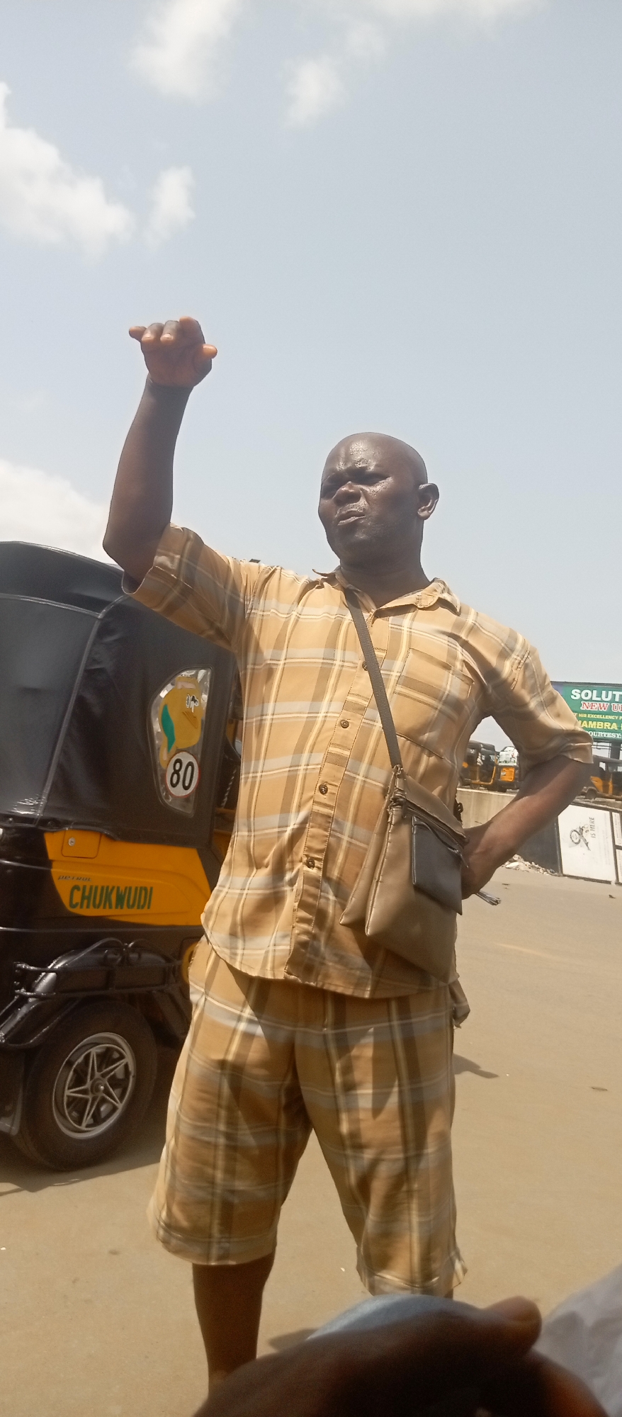The image shows a bald man with dark skin standing on a street in Onitsha, Nigeria. He is wearing a tan and grey plaid short sleeve shirt and matching shorts, with a tan shoulder bag. His left arm is raised in a fist, and his right hand is on his hip. Behind him is a black and yellow auto rickshaw, with the name "CHUKWUDI" and the number "80" visible. In the background is a sign that reads "SOLUTION" and "NEW UPD", likely referencing a political candidate or initiative. The sky is bright and lightly clouded. The scene suggests a possible demonstration or political rally.