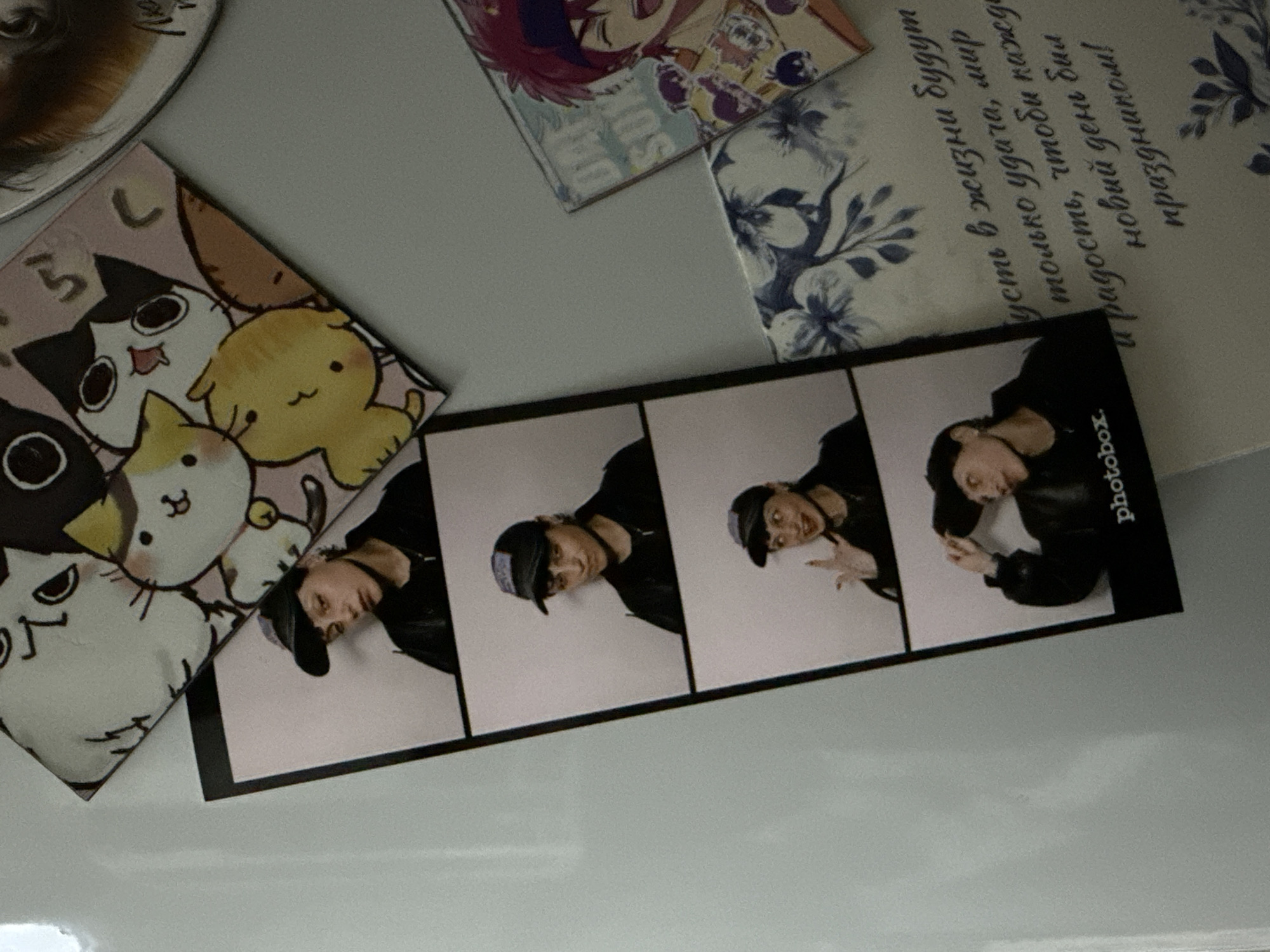 The image appears to be a close-up shot of a collection of items on a white surface. The items include: a round object (possibly a coaster or magnet) with a brown and white pattern, a square-shaped object with anime-style illustrations of cats and a yellow creature, a square-shaped object with an anime-style character, a square-shaped object with blue floral designs and Russian text, and a strip of photobooth pictures. The photobooth strip contains four images of a person wearing a black jacket and a baseball cap, posing in different ways. The word "photobox" is printed on the side of the photobooth strip. The text in the Russian text is "пусть в жизни будут только удача, мир и радость, чтобы каждый новый день был праздником" which translates to "May there only be luck, peace and joy in life, so that every new day is a holiday!". There is no evidence to suggest the location is Saint Petersburg, Russia.