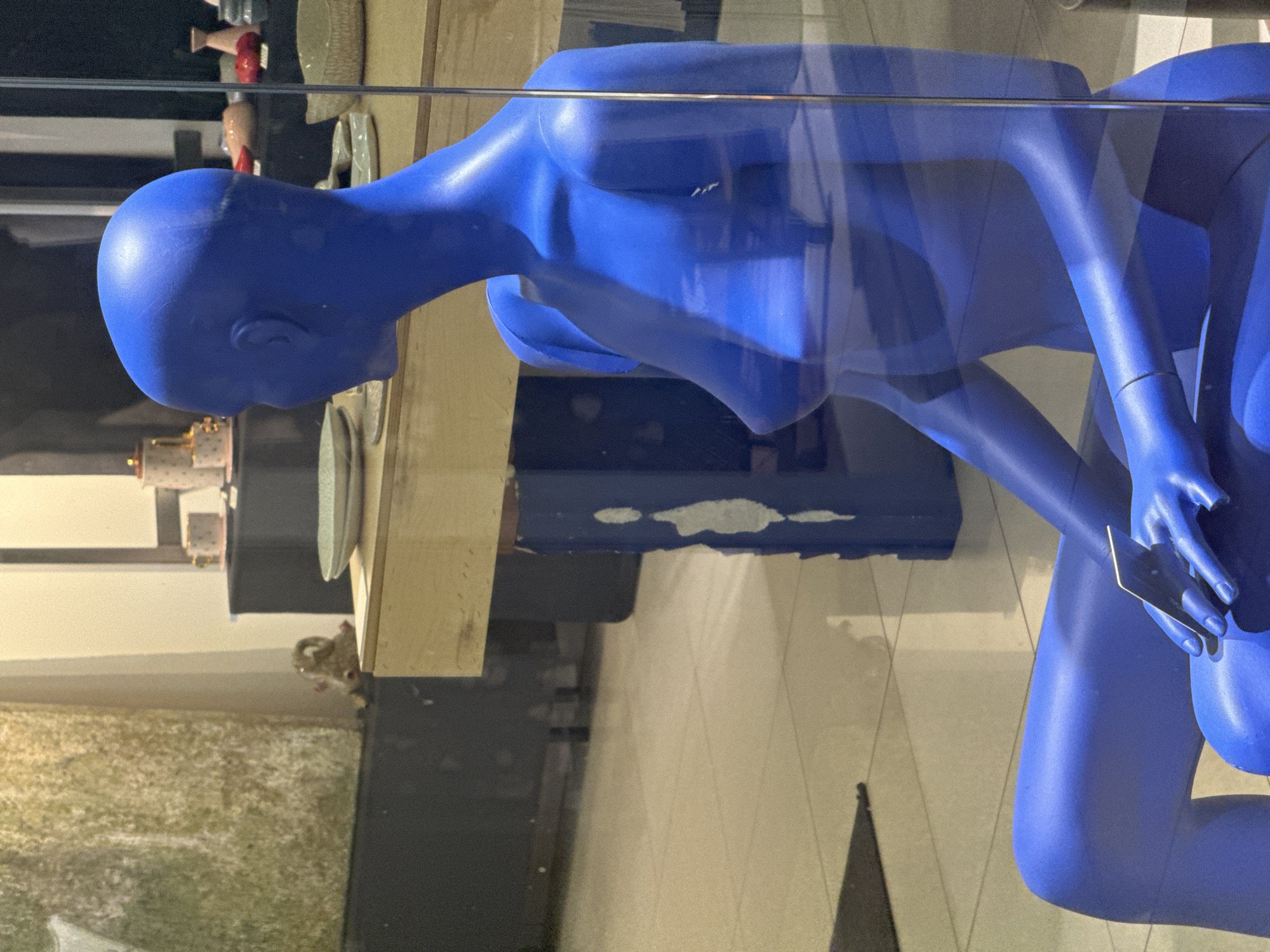 The media file presents a vibrant, matte blue, featureless mannequin displayed behind a clear glass barrier, suggesting a store window or exhibition. The mannequin, appearing female due to its form, is positioned in a crouched or seated pose, with its head turned to the left and looking downwards. Its right hand is delicately holding a small, dark, rectangular object, possibly a credit card or a miniature smartphone, between its thumb and fingers.
The clear barrier creates numerous reflections, superimposing elements of the surrounding environment onto the mannequin and the background. Visible through the glass, behind the mannequin, is a display area featuring a wooden shelf or counter. On this shelf, several small decorative items can be discerned, including a reddish-colored figure and what appears to be a small animal figurine (possibly a squirrel) peeking from behind another object. Further back, a dark background with some lighter patches (possibly flaking paint on a blue surface) is present.
Reflected on the glass, and appearing to overlap the mannequin and background, are patterns suggesting a tiled or textured floor and indistinct shapes from the broader space. A prominent horizontal line, likely the edge of a display case or shelf, runs across the image. The mannequin itself has visible seams on its head and limbs, typical of its construction. The lighting appears artificial, indicating an indoor setting. No discernible text is visible. The scene is located in Krasnoyarsk, Russia.