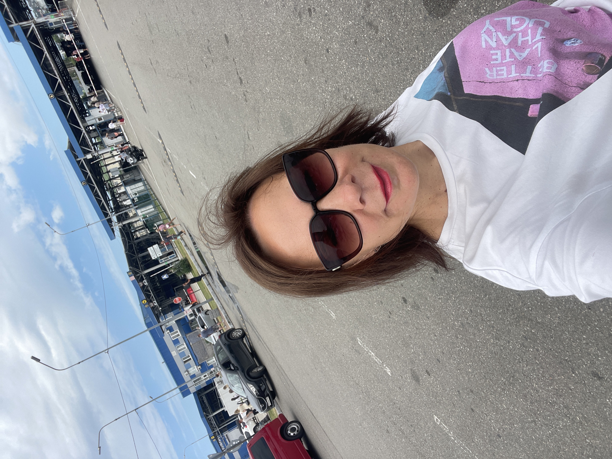 A woman with shoulder-length brown hair and dark sunglasses takes a selfie outdoors. She is wearing a white t-shirt with a graphic design that reads "BETTER LATE THAN UGLY" in pink lettering. The background shows a border crossing with multiple lanes for vehicles, and a long, blue and white building with an overhang supported by pillars. There are several cars, including a black SUV and a red van, and people walking around. The sky is cloudy. The location is identified as a border crossing in the city of Sirius, Russia.