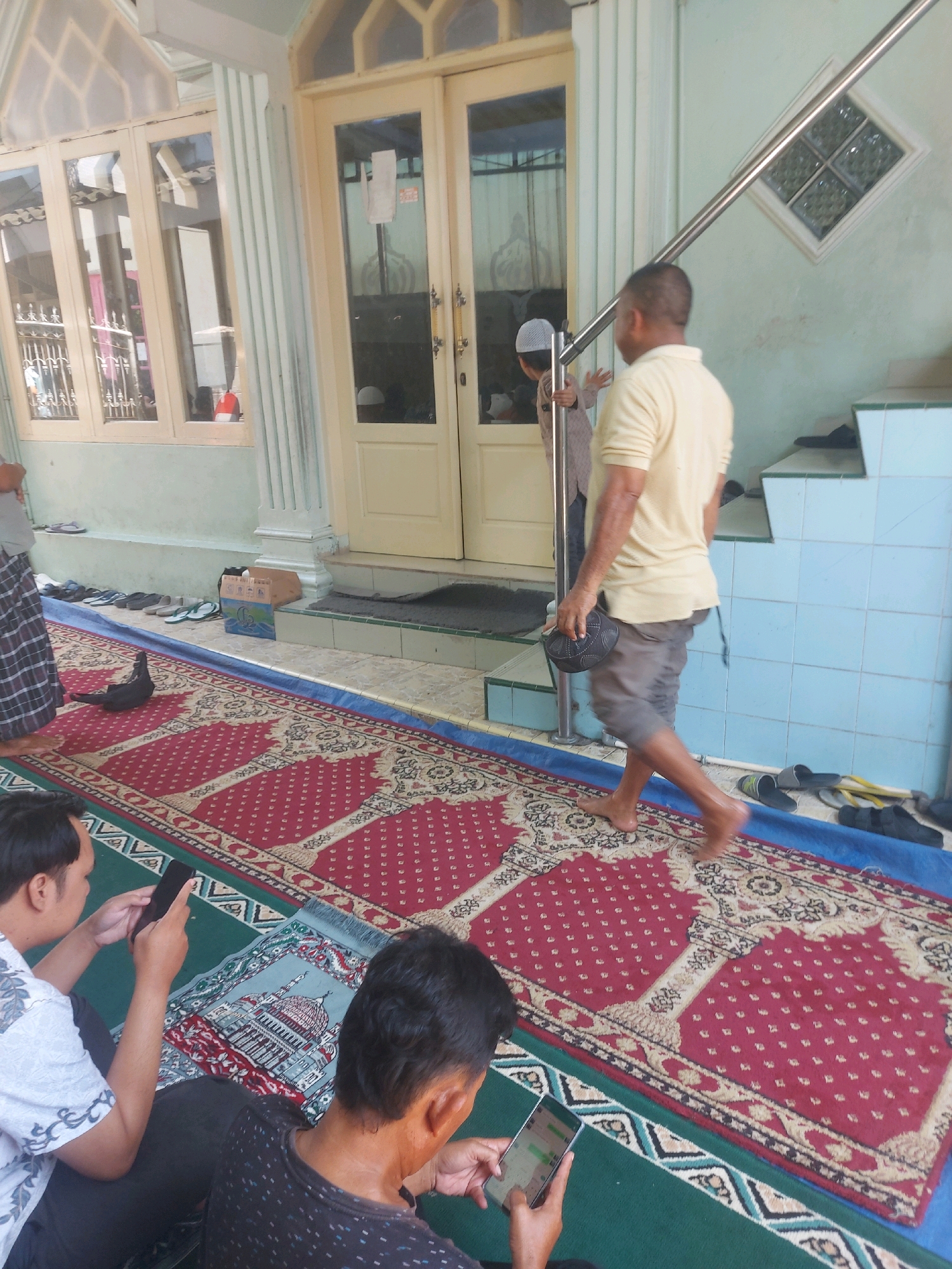The image depicts the exterior of a mosque in Makassar, Indonesia, with several individuals present. A man in a yellow t-shirt and grey shorts is walking away from the camera, holding a dark cap. To his left, a child wearing a white peci (cap) is visible near a doorway. In the foreground, two men are seated on prayer mats, one looking at his mobile phone and the other holding a tablet. The mosque features large glass doors, windows with decorative grills, and a light green painted wall. A long, red prayer rug with a traditional pattern is laid out on the ground, extending towards the entrance. Various pairs of sandals and shoes are placed neatly along a blue mat near the mosque entrance. The overall scene suggests a communal gathering at the mosque, likely for prayer or religious activity.