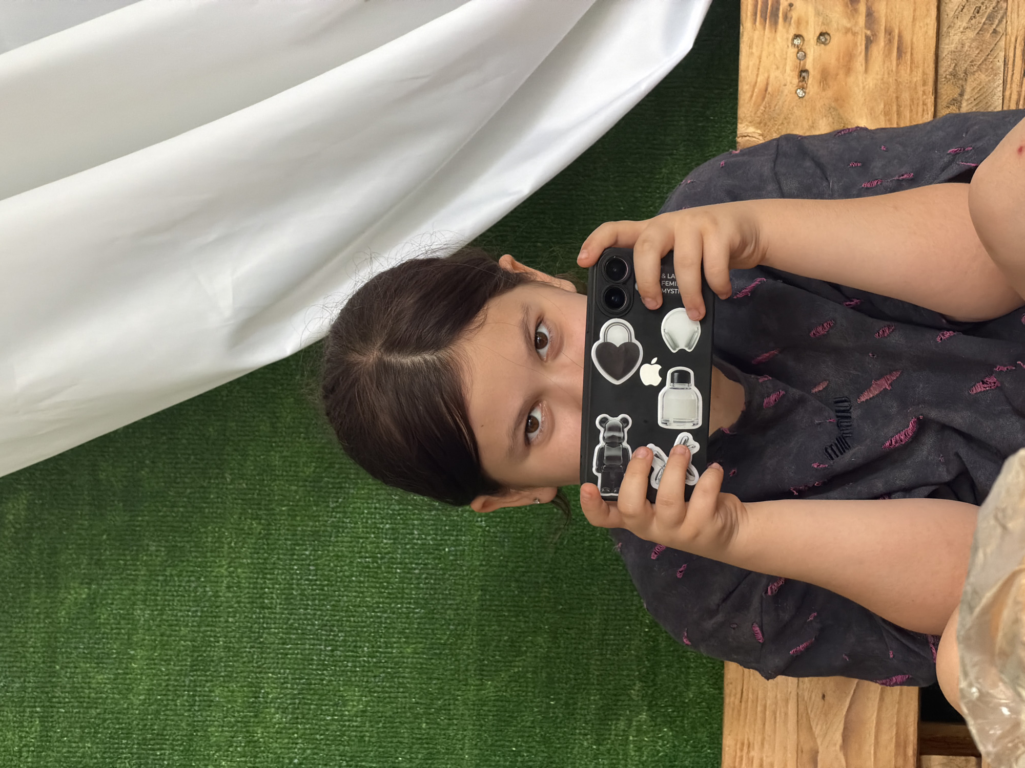 An eye-level, medium shot captures a young girl in Tuapse, Russia, holding a black smartphone horizontally in front of her face. She is seated on a wooden bench, with her dark hair pulled back. The phone case is adorned with several white stickers, including a heart, a bear, and an apple logo. Behind her, a textured green surface, possibly artificial grass, contrasts with the draped white fabric to her right. The girl is wearing a dark grey t-shirt with rips and pink threads visible. The scene appears to be outdoors, possibly in a park or recreational area.
