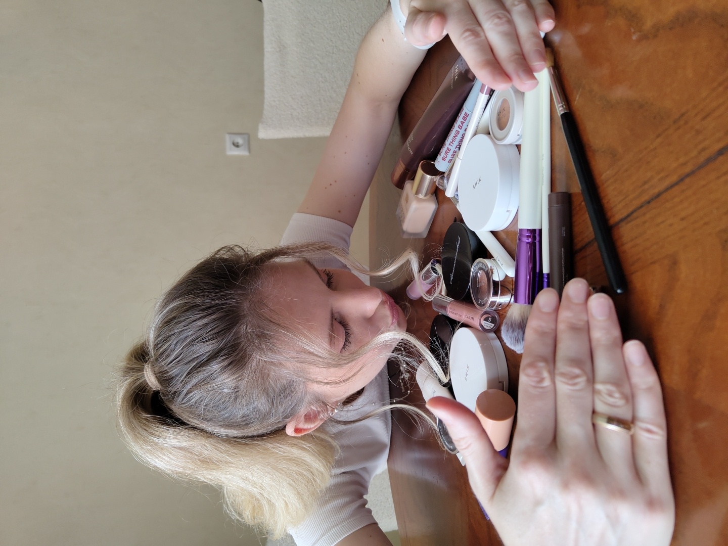 A blonde woman with her hair in a high ponytail is leaning over a wooden table filled with makeup. She is wearing a white shirt and has a ring on her left ring finger. Her hands are spread out on either side of the makeup. The makeup includes various items such as foundations, powders, lipsticks, brushes, and pencils. The background appears to be an indoor setting with a wall and a chair visible. The scene is well-lit, suggesting it is daytime. The image does not provide any specific information to determine its location within Krasnodar, Russia.