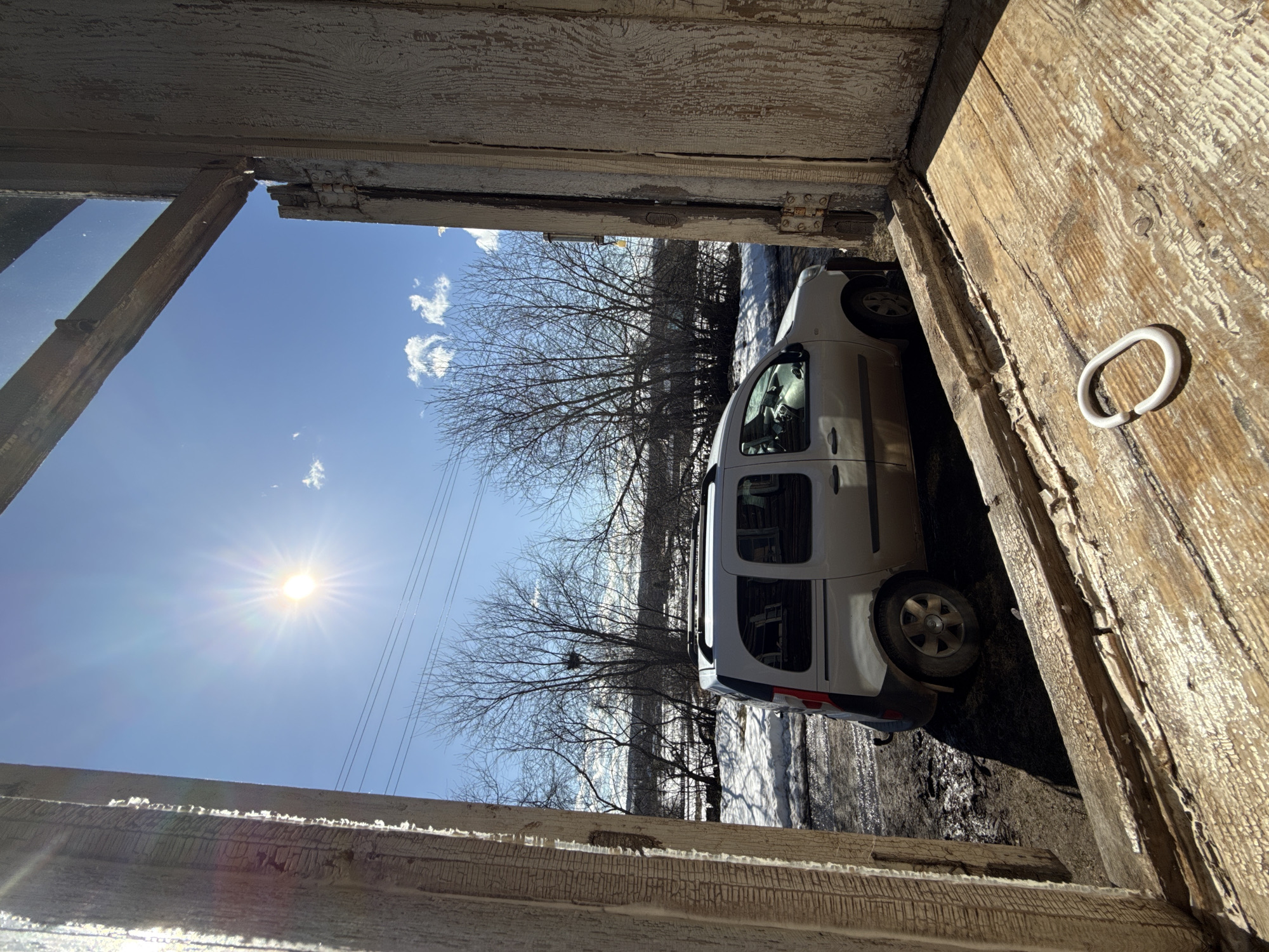 The image depicts a white van parked outside, viewed from inside a dilapidated wooden structure, possibly a shed or outbuilding. The exterior scene includes bare trees, snow, and utility lines against a bright blue sky with the sun visible. The wooden structure has peeling paint and a metal hook is attached to the right side of the frame. The van is parked close to the structure, partially obscuring the view of the trees and snow. The exact location is Nizhnyaya Chermoda, Russia.