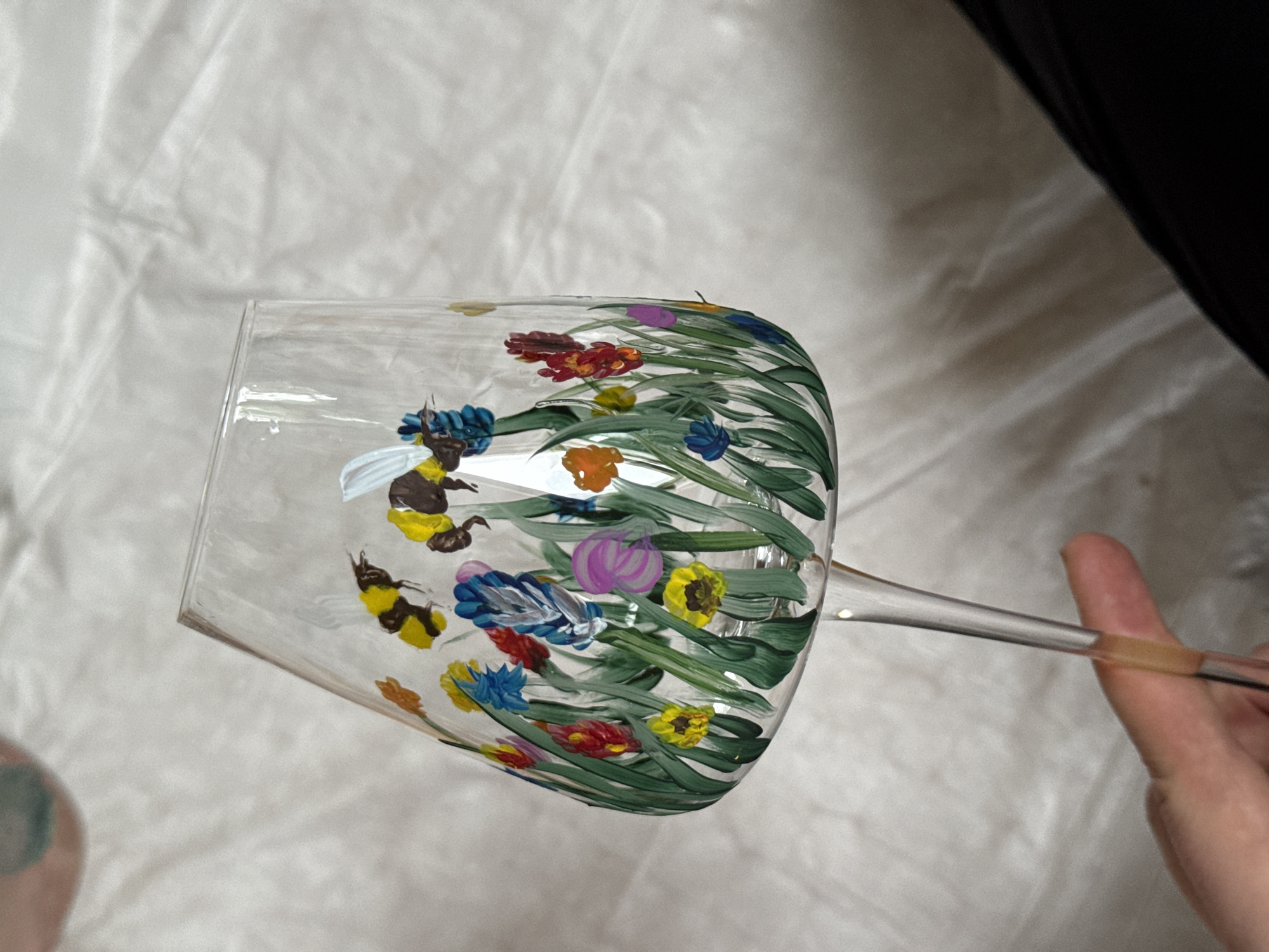 A close-up shot shows a hand holding a wine glass decorated with painted flowers and bees. The background consists of wrinkled white fabric and a dark, undefined area. The subject being painted is in an indoor setting. There is no indication of the location or existence of Sapronovo, Russia.