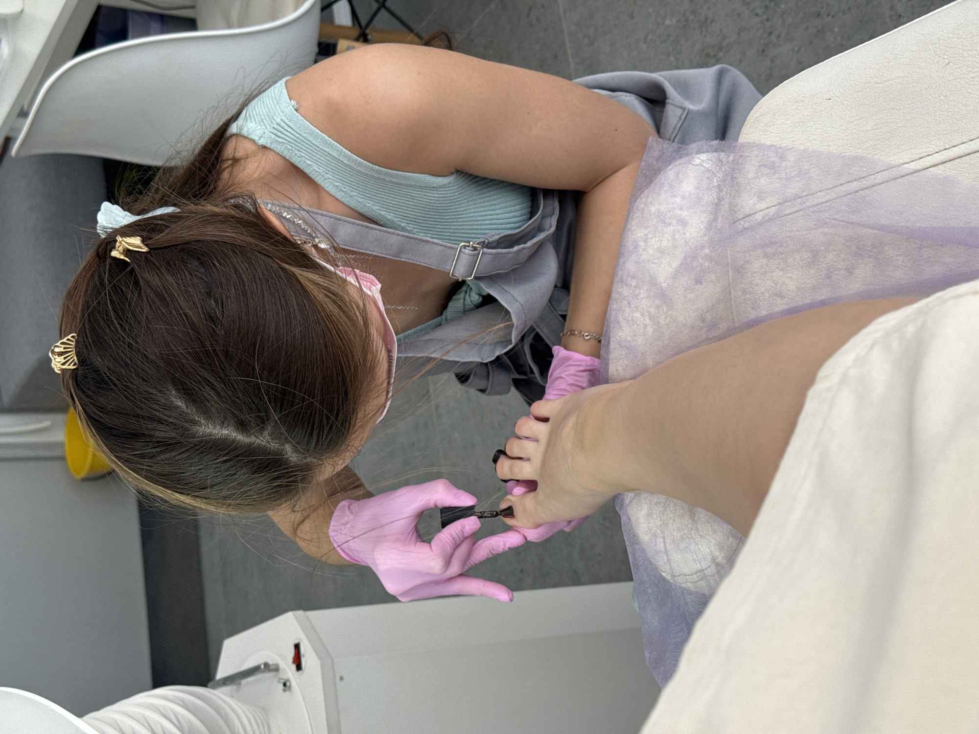 A woman is painting a person's toenails. The woman is wearing pink gloves and a light blue tank top under a gray overall dress. She has brown hair with gold butterfly clips. The person whose toenails she is painting is sitting in a chair covered with a light purple fabric. The scene is indoors, with a pedicure machine visible in the foreground and other salon furniture in the background. The location is a nail salon, potentially in Izhevsk, Russia.