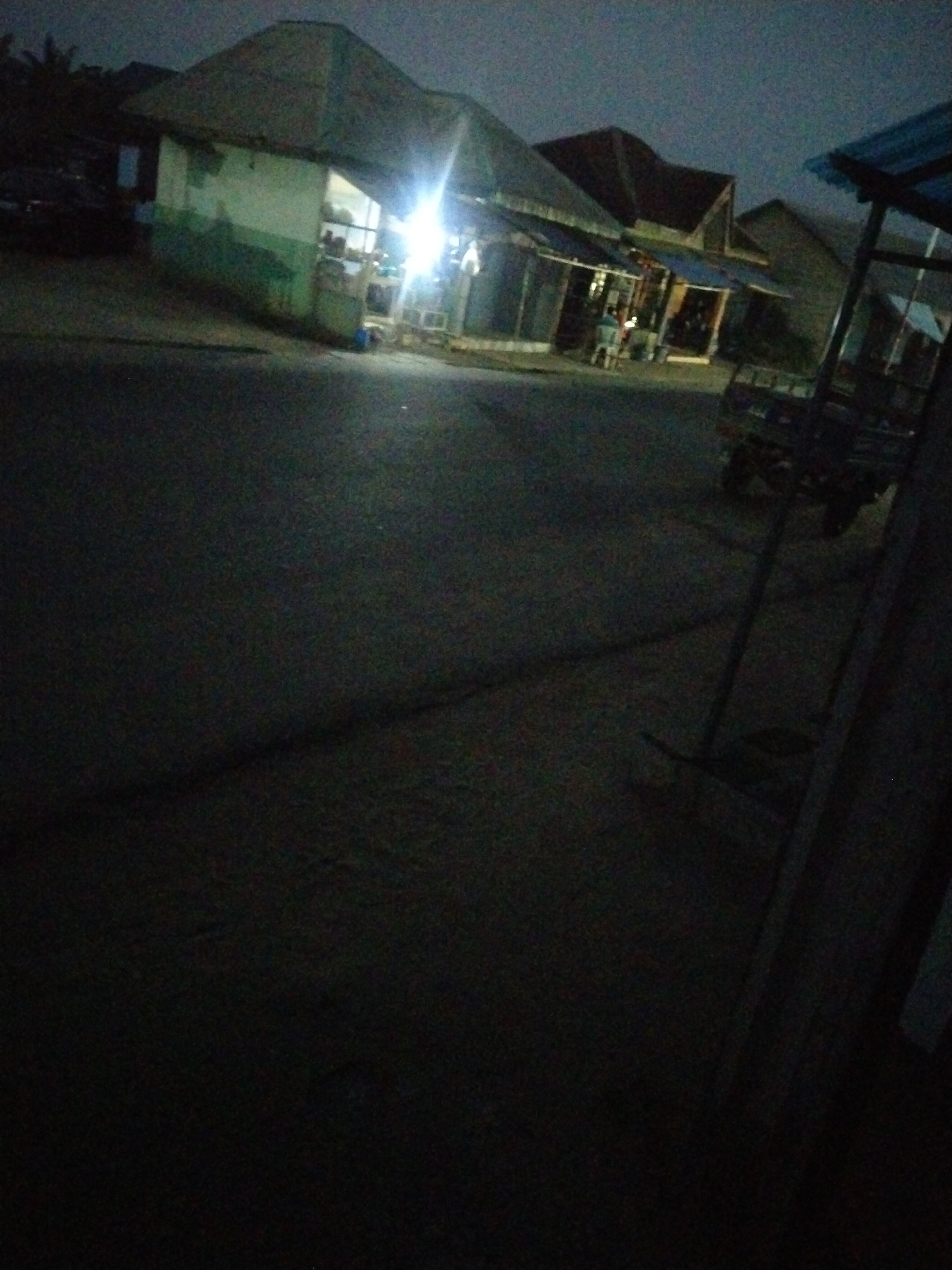 The image depicts a street scene in Uyo, Nigeria at dusk. The street is dark and mostly deserted. Buildings line the street, including a small shop illuminated by a bright light. A vehicle, possibly a three-wheeled auto rickshaw, is parked near the shop. The overall environment suggests a quiet residential or commercial area at twilight. There are 2 people standing outside the shop.