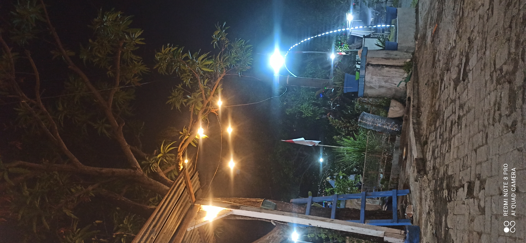 The image depicts an outdoor scene at night in Sedati Agung, Indonesia, likely a public space or a small establishment with outdoor seating. A large tree with visible branches and leaves dominates the upper portion of the frame, its foliage dark against the night sky. A corrugated metal roof structure, supported by wooden beams, is visible on the left, with string lights illuminating the area. These lights, some round and others small bulbs, are strung across branches and overhead. A blue ladder leans against a wooden post. In the mid-ground, an Indonesian flag, red and white, stands on a pole. Various plants and foliage are present, some in blue planters. A concrete structure serves as a planter or seating area. A bright, intense light source is positioned on the right side of the image, casting a strong glow and creating lens flare effects. Below this light, a string of smaller white lights forms a curved arc. The ground in the foreground is paved with dark bricks or cobblestones, appearing damp. The overall atmosphere is dimly lit and appears to be a nighttime setting, possibly a garden or a roadside eatery.
